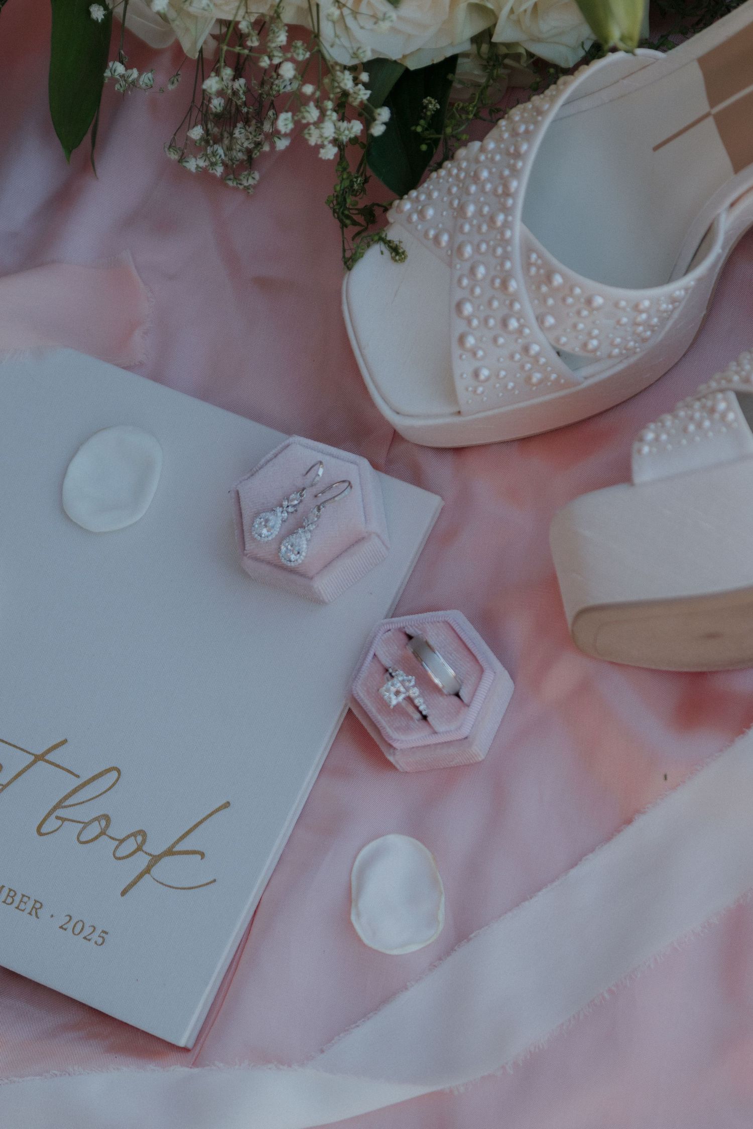 Wedding details including pearl-adorned shoes, rings in hexagonal boxes, and a thank you note on pink silk fabric.