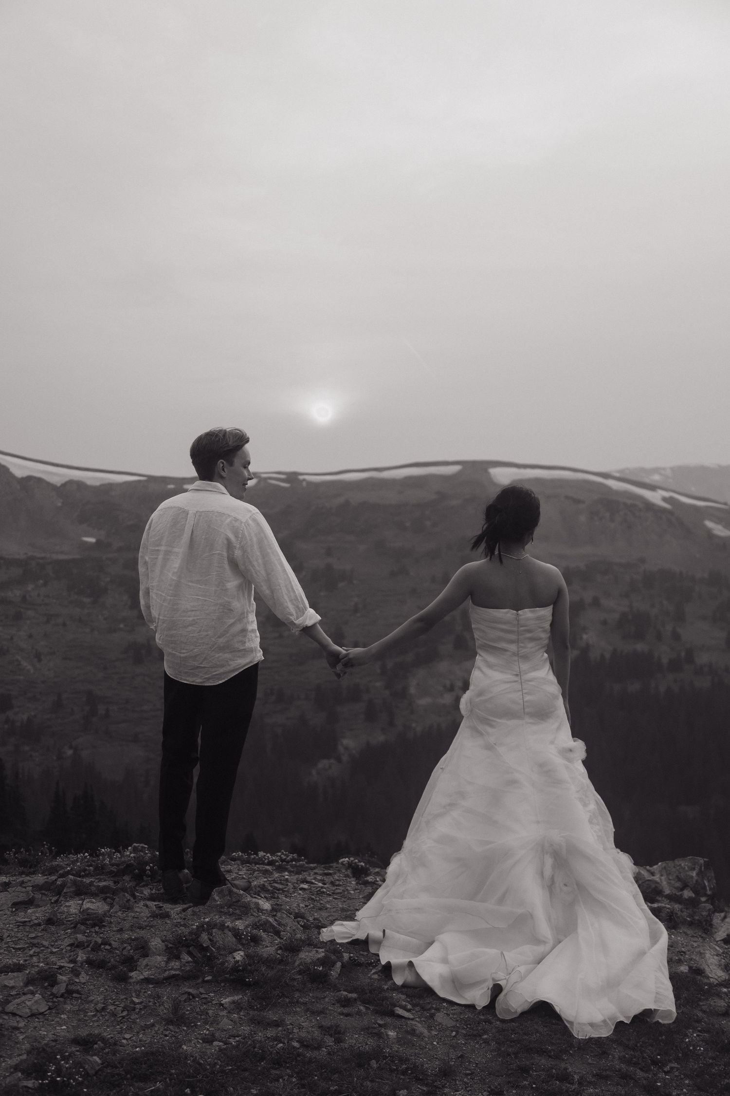 Black and white photo of bride and groom holding hands while gazing at a misty mountain sunset.