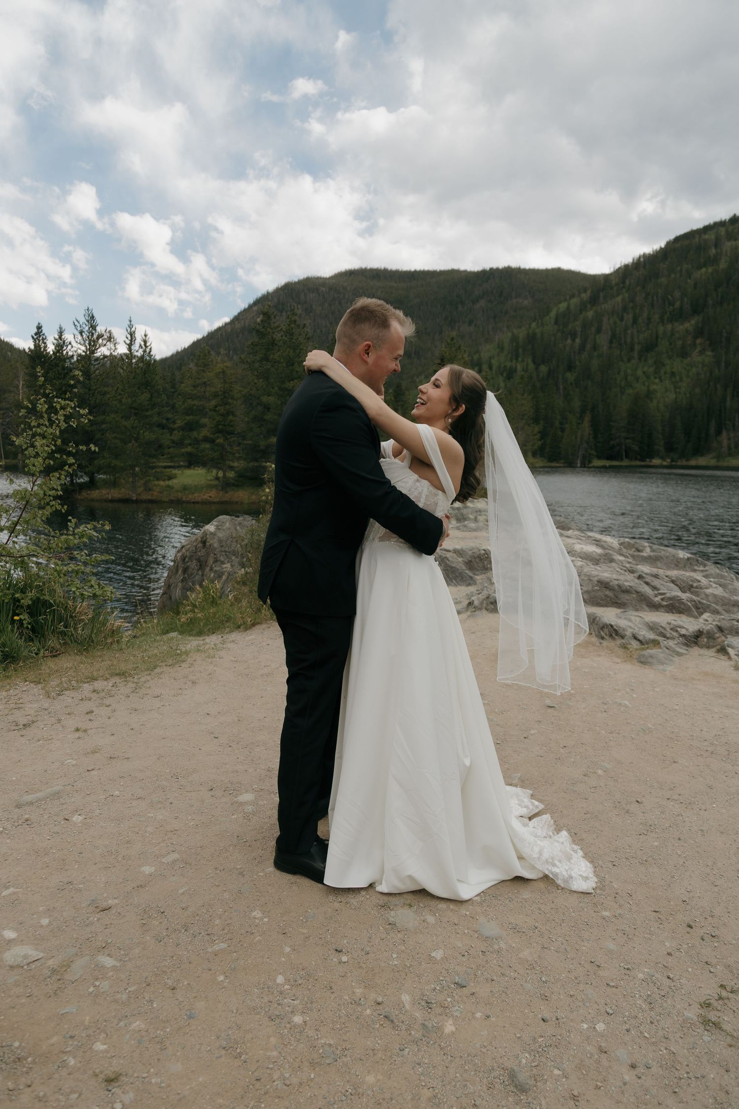 Bride and groom embrace tenderly by a mountain lake, with pine trees and cloudy sky in the background.