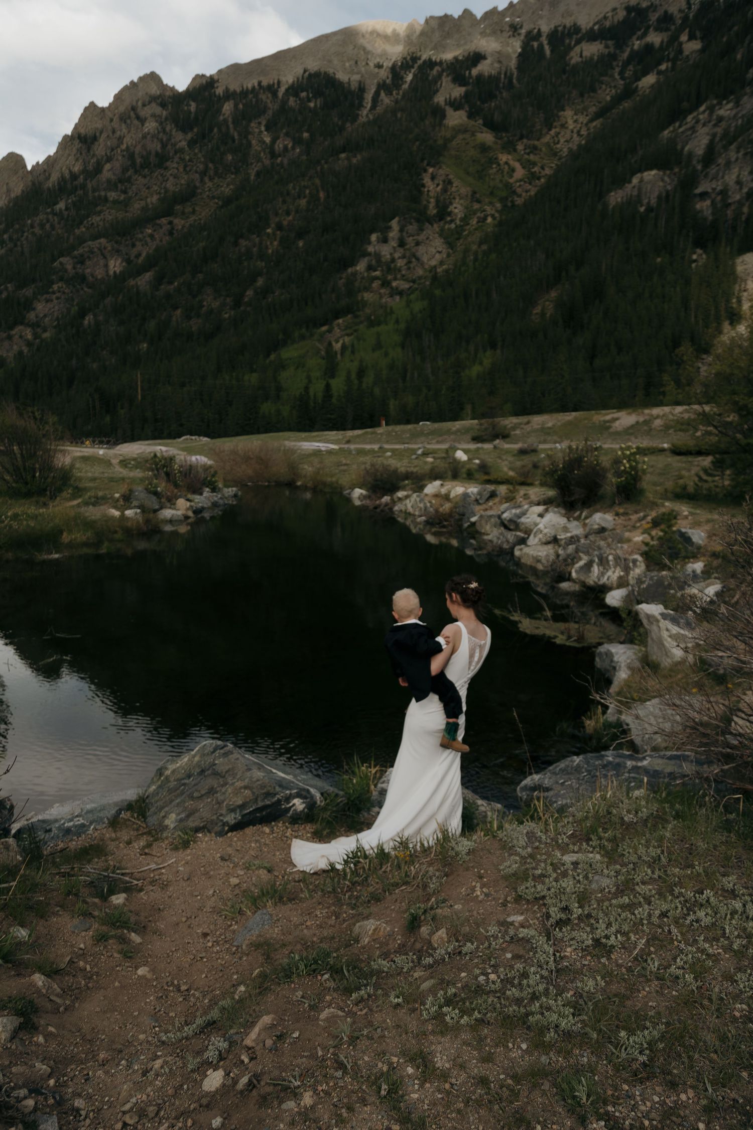 Bride and groom stand together on rocky shore beside dark mountain lake during outdoor elopement ceremony.