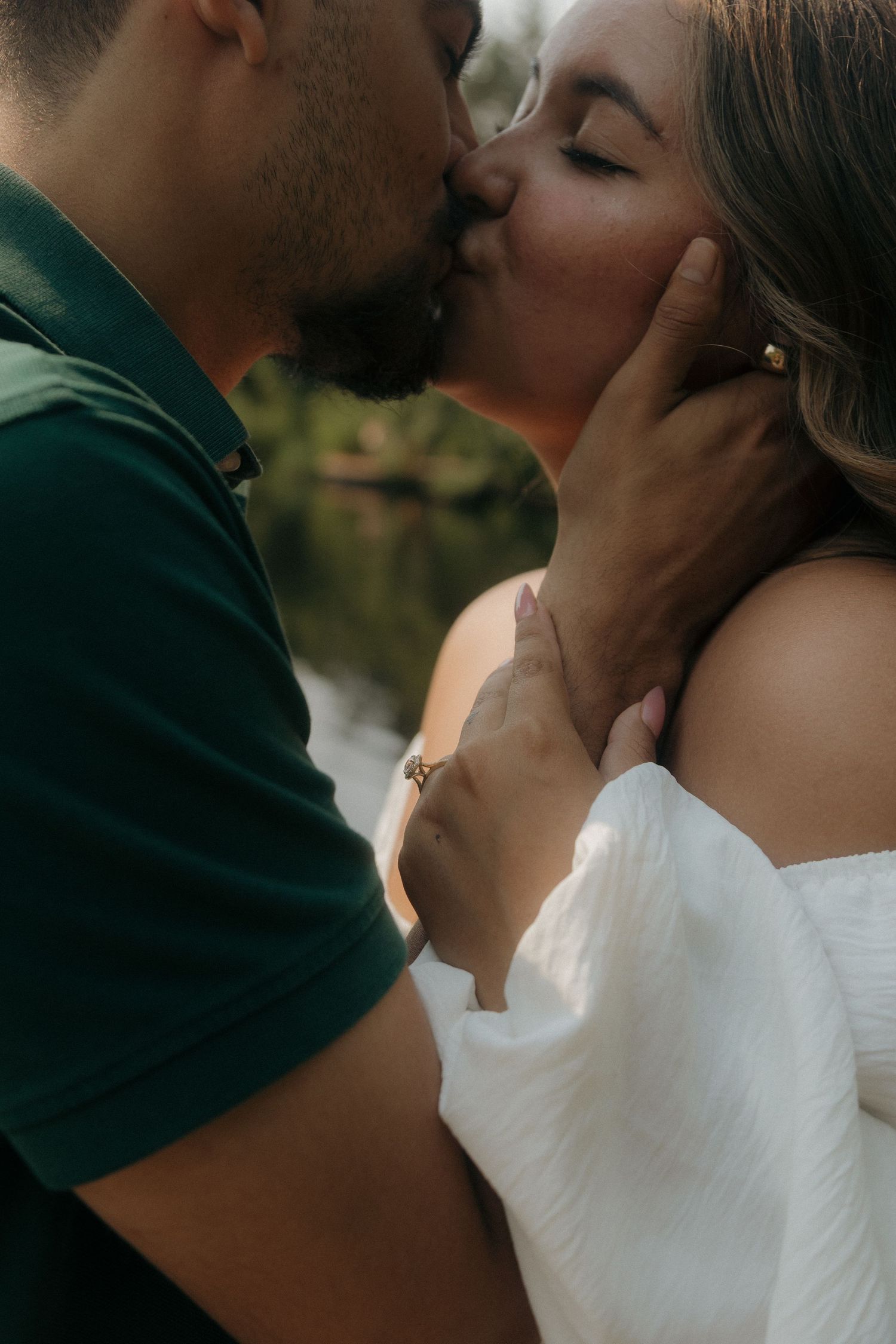 Couple sharing a romantic kiss outdoors, man in green polo gently holding woman's face in white dress.