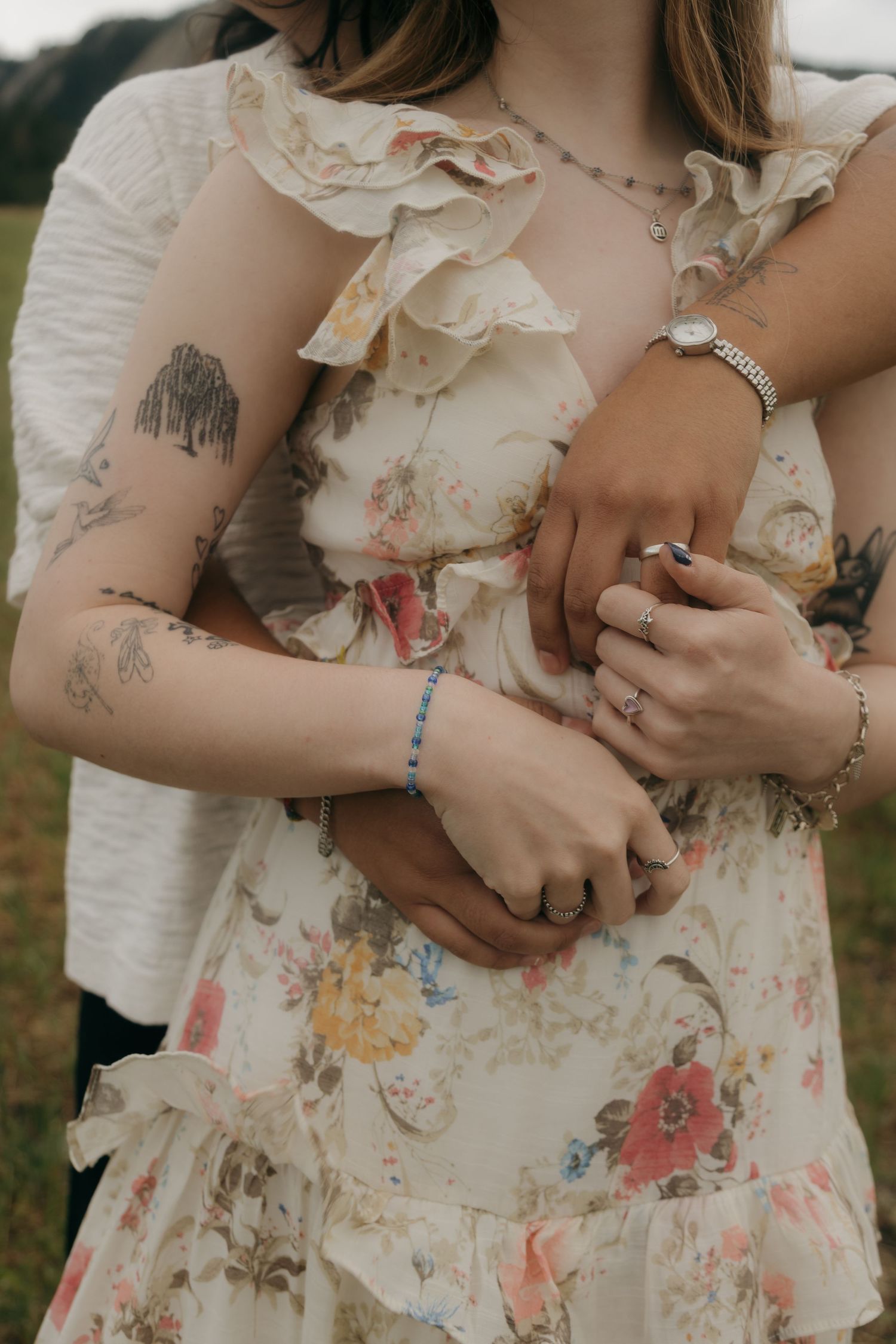 Two people embracing, woman in floral ruffle dress with tattoos, both wearing rings and bracelets outdoors.