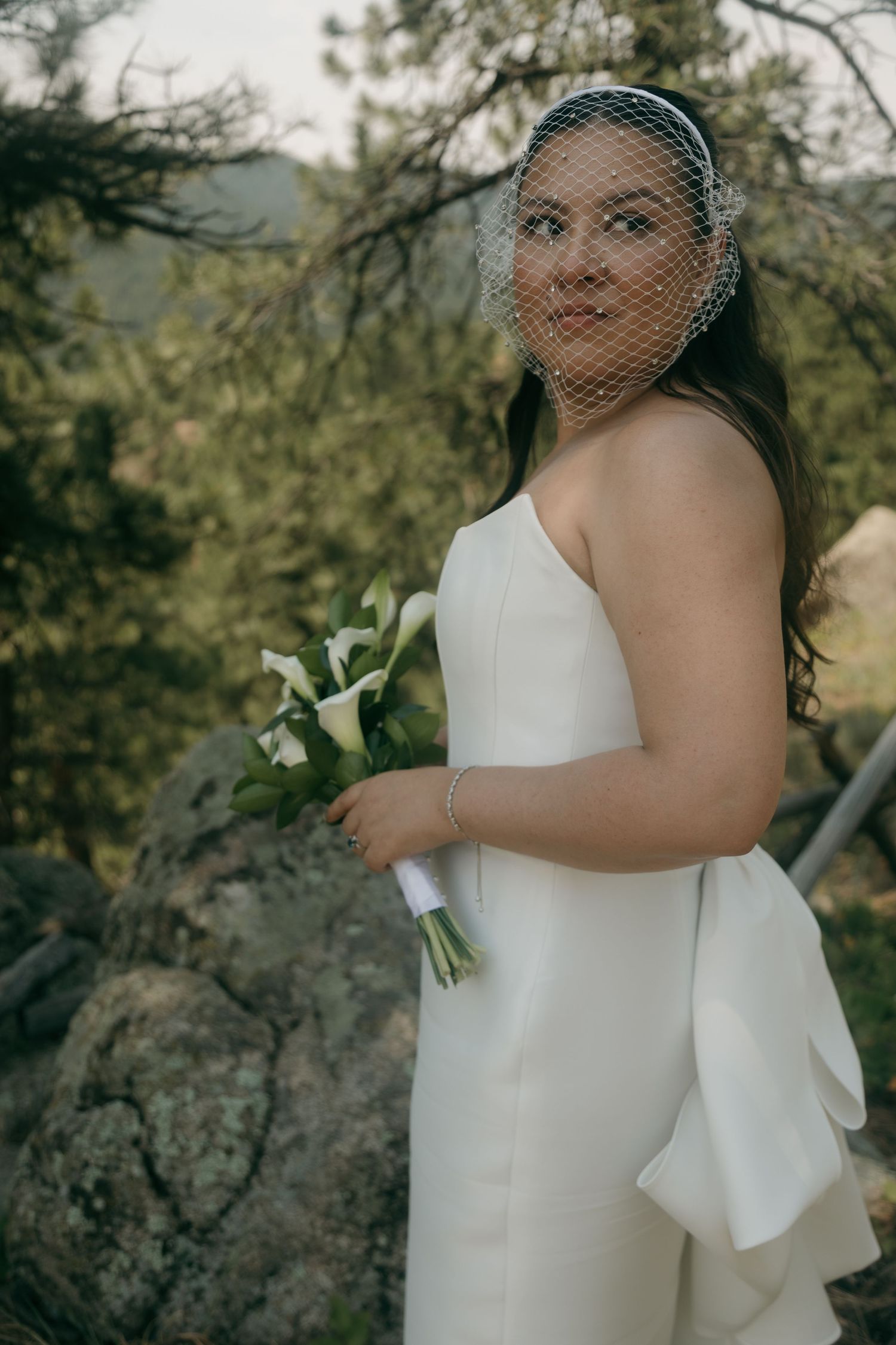 Bride in white strapless gown and birdcage veil holding calla lily bouquet among rocky scenery.