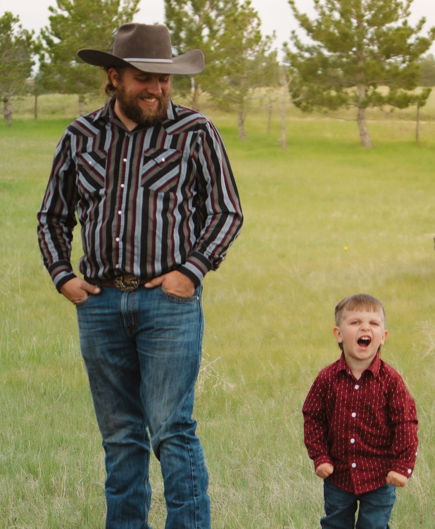 A cowboy in striped shirt and child in red button-up share a joyful moment outdoors.
