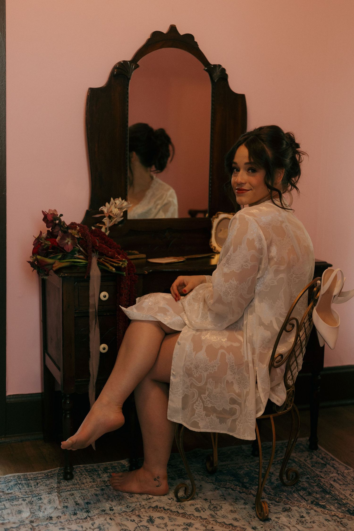 Woman in white robe sits at vintage vanity mirror with elegant updo hairstyle.