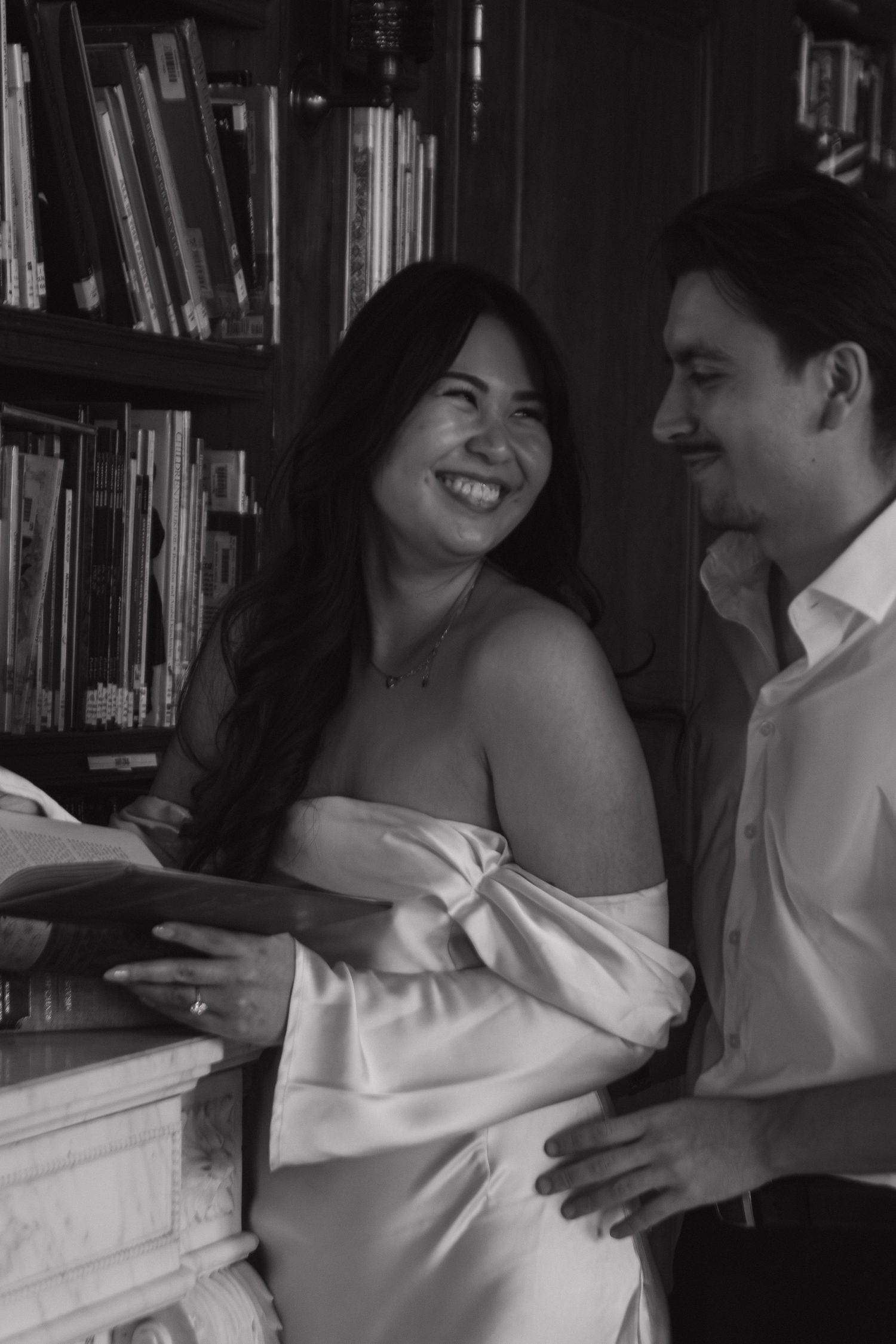 A romantic black and white photo of a couple sharing a joyful moment in front of a bookshelf.