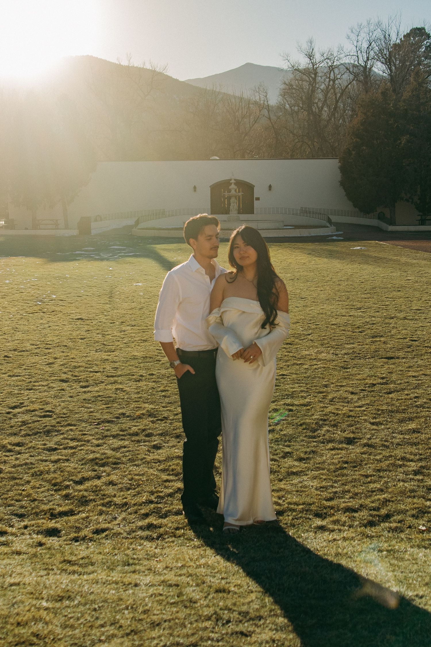 A couple stands together on a lawn at golden hour with mountains and architecture in the background.