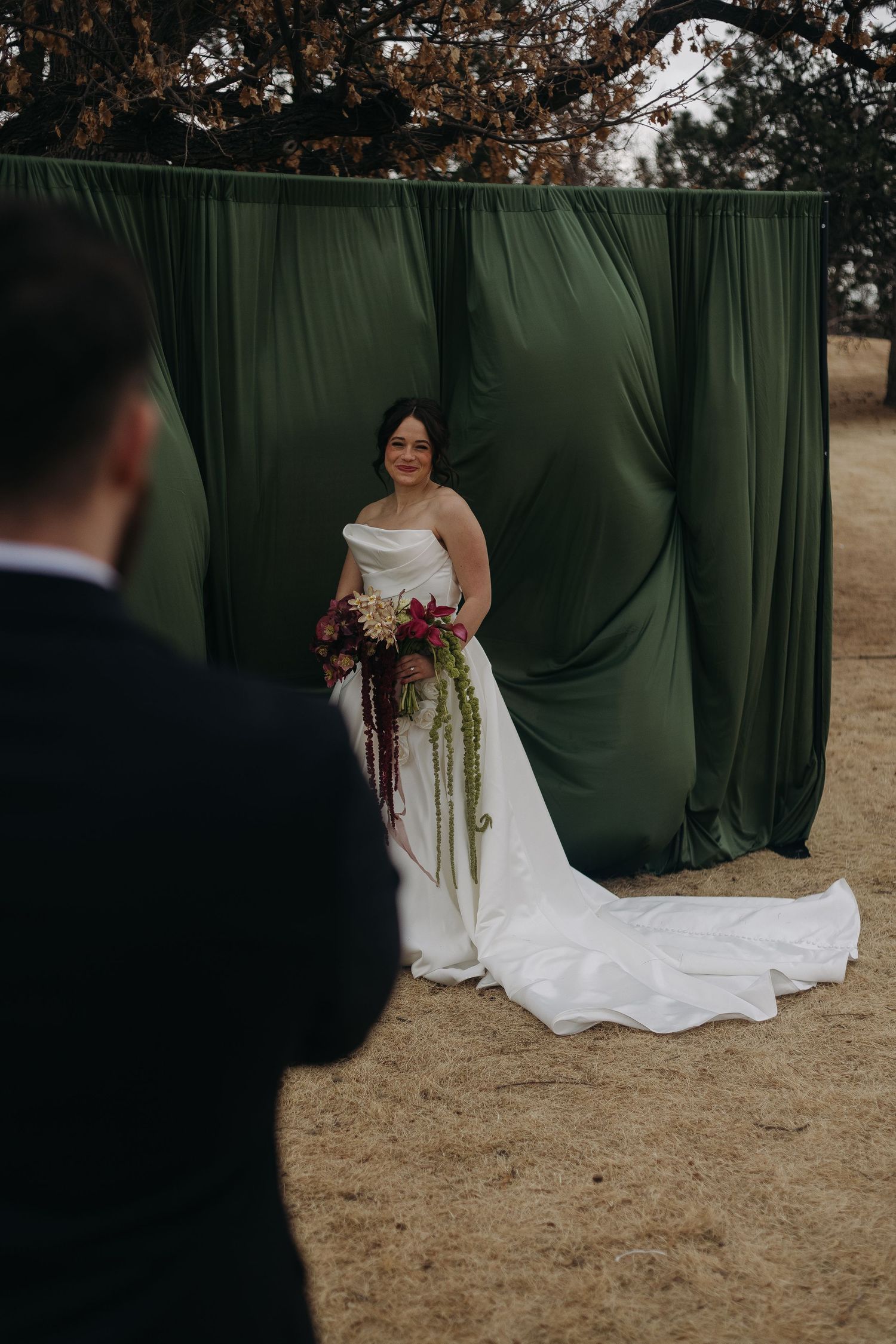 Elegant white wedding dress with a flowing train photographed against a dark green backdrop outdoors.