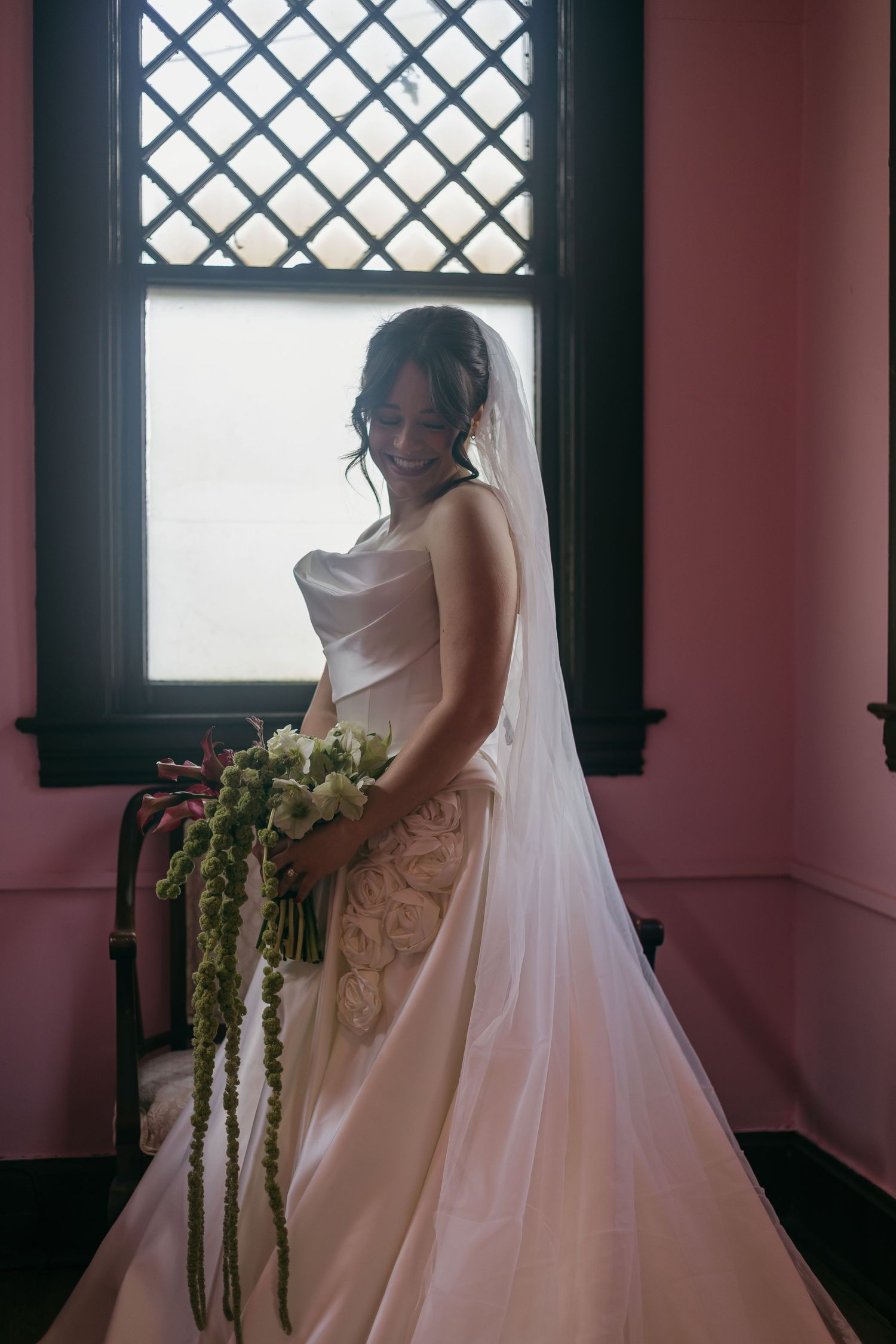 A bride in a flowing white wedding dress stands by a window with a cascading green floral bouquet.