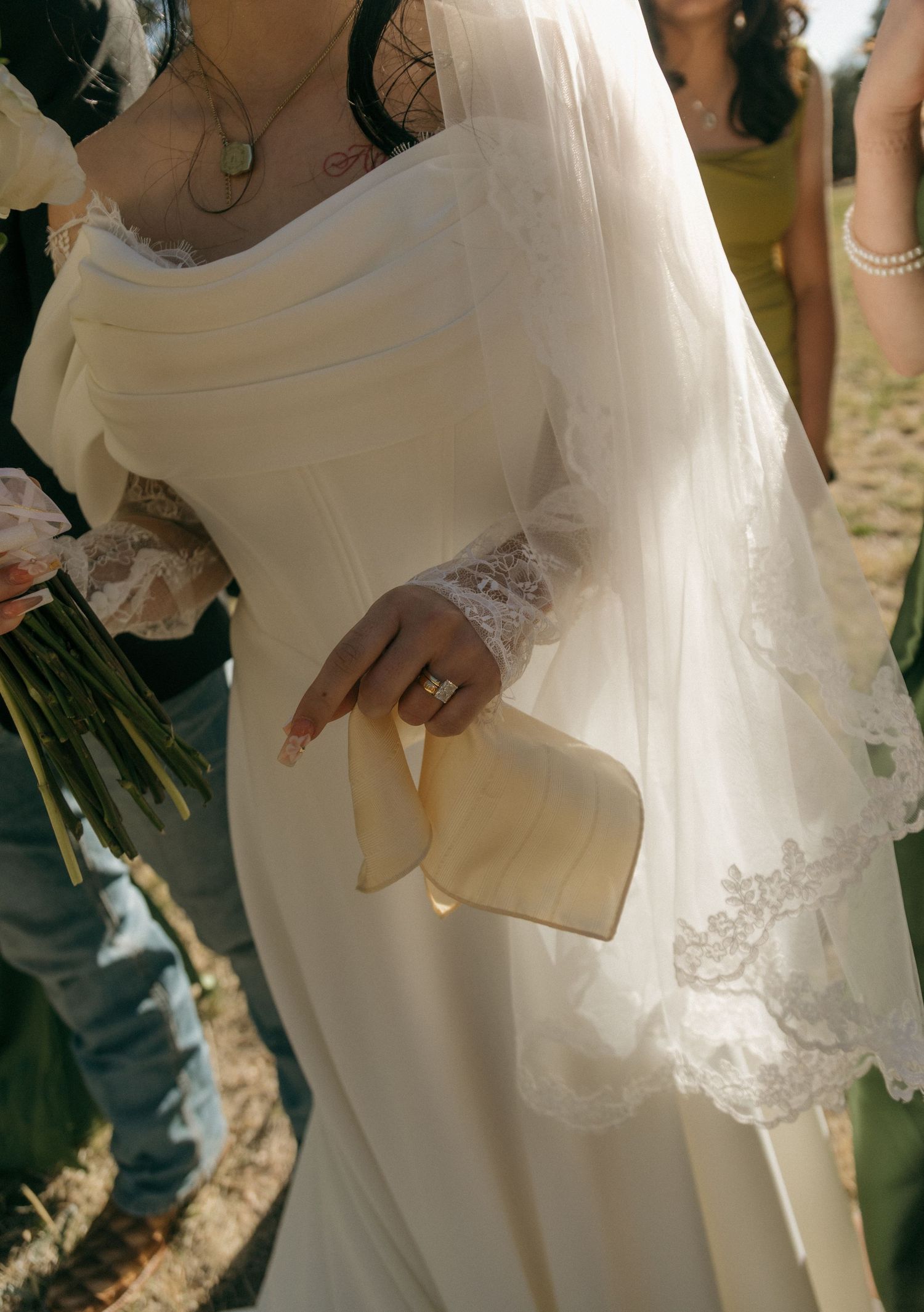 Bride in ivory lace-sleeved wedding gown holding a ribbon, standing outdoors with bridesmaids in olive dresses.