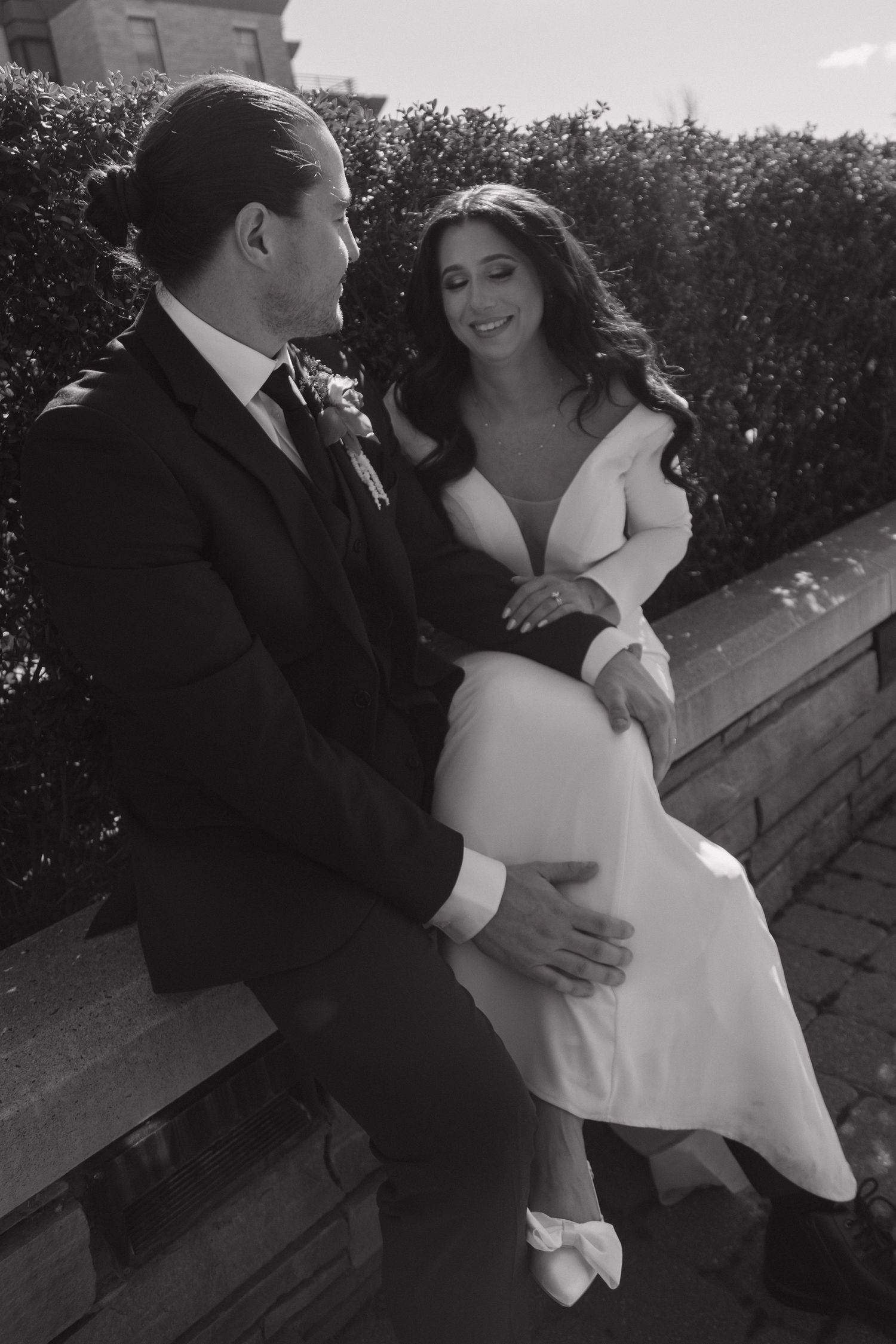 Black and white photo of bride and groom sitting together on an outdoor stone ledge.