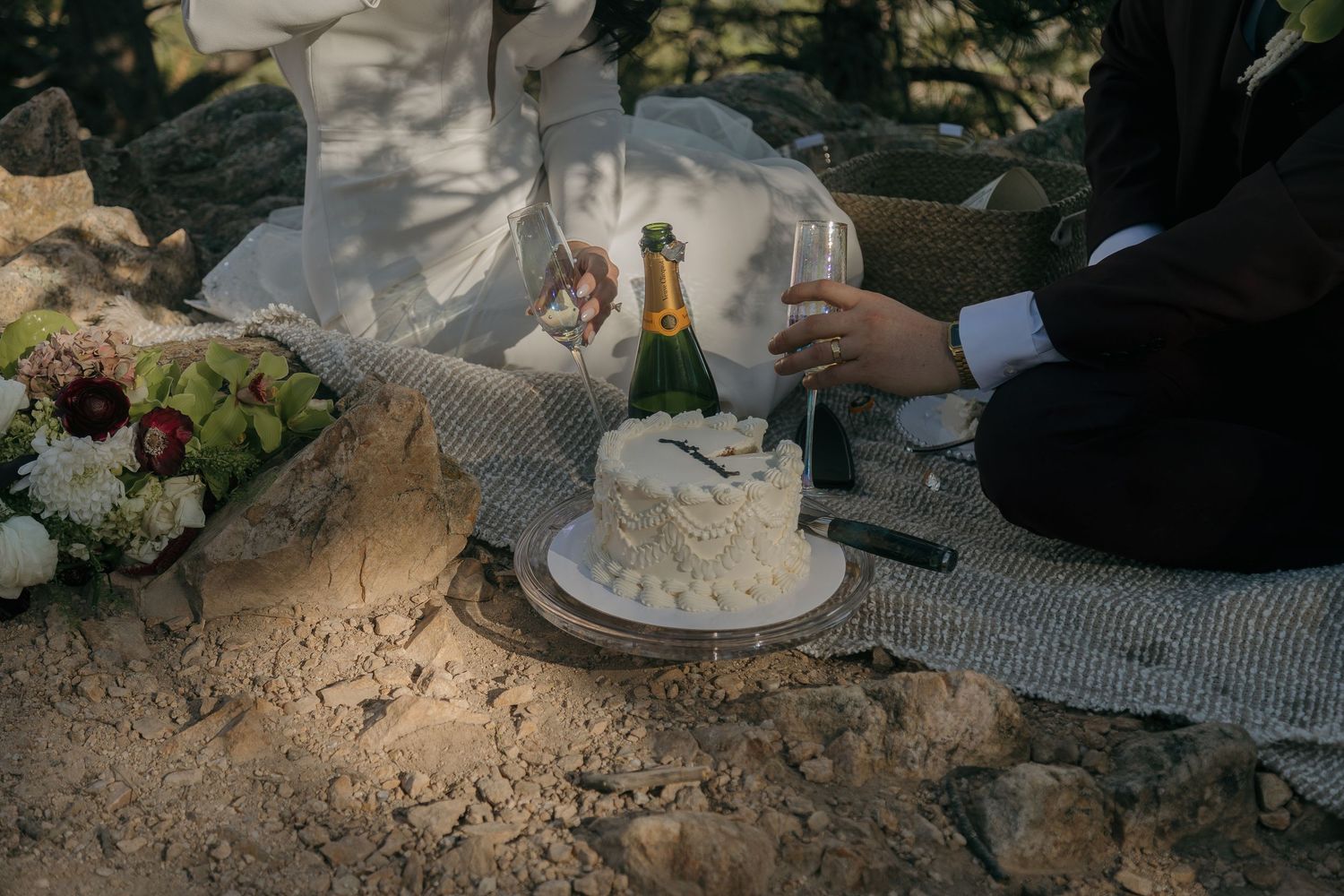 Newlyweds cut a white wedding cake on rocks beside champagne and a floral bouquet.