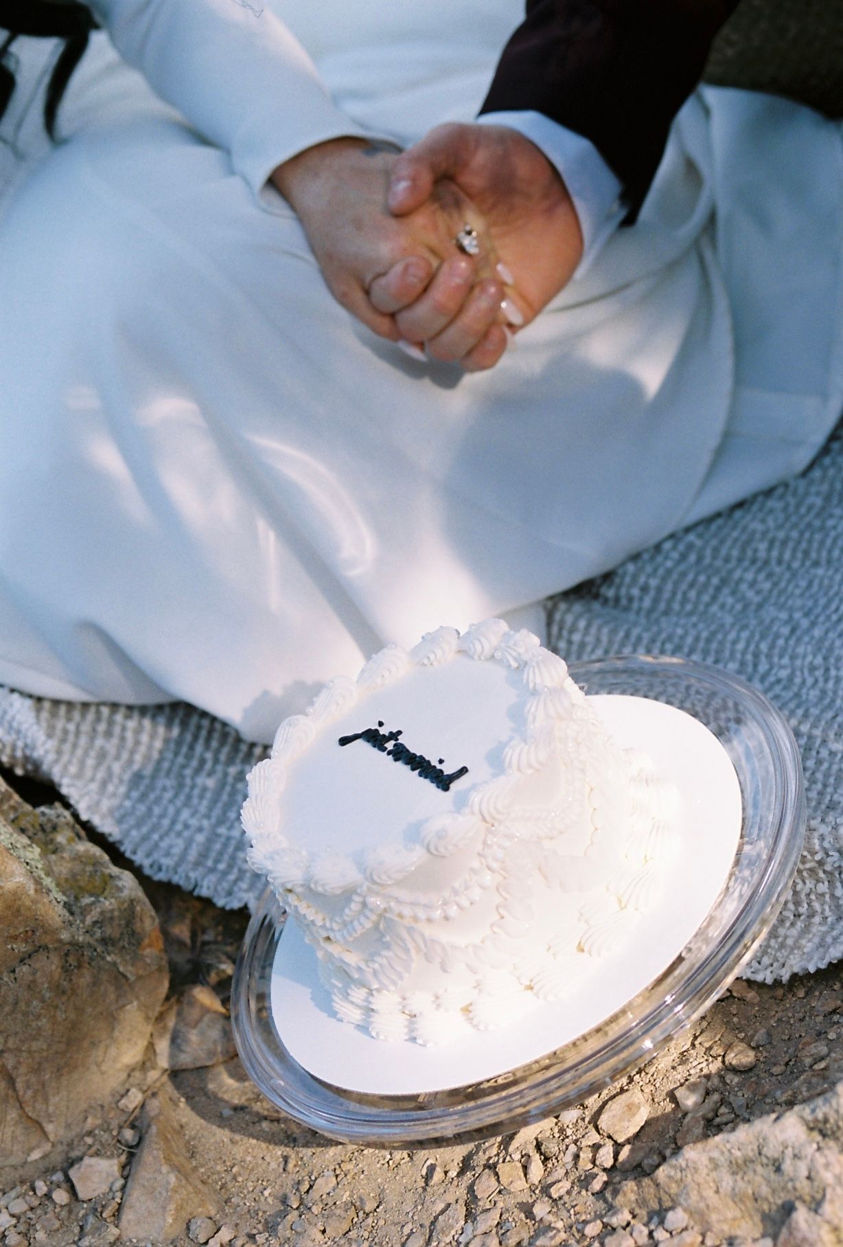 White heart-shaped wedding cake with decorative frosting and personalized topper placed on rocky outdoor surface.