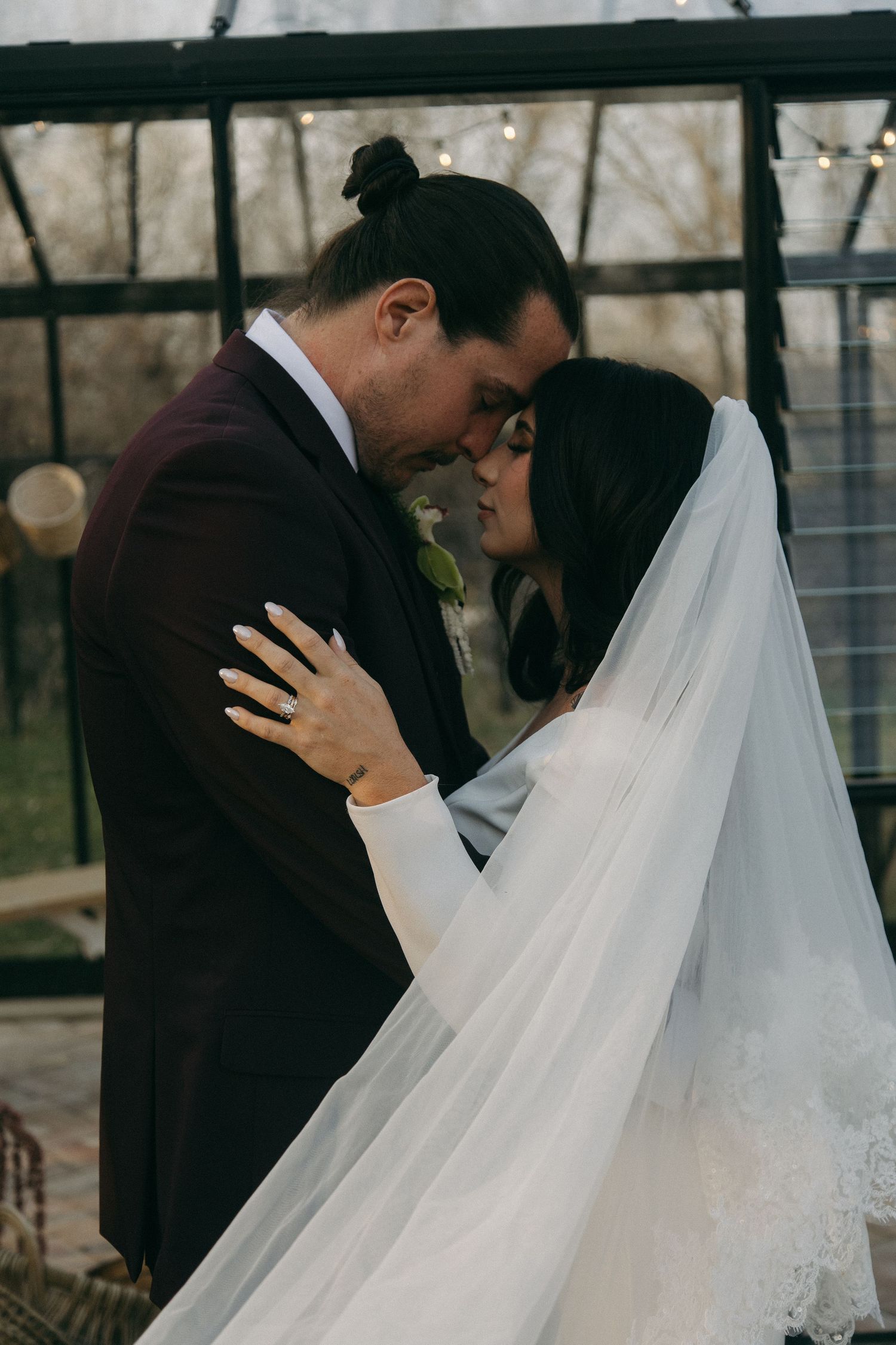 Bride and groom sharing intimate kiss under flowing veil in front of glass greenhouse structure.