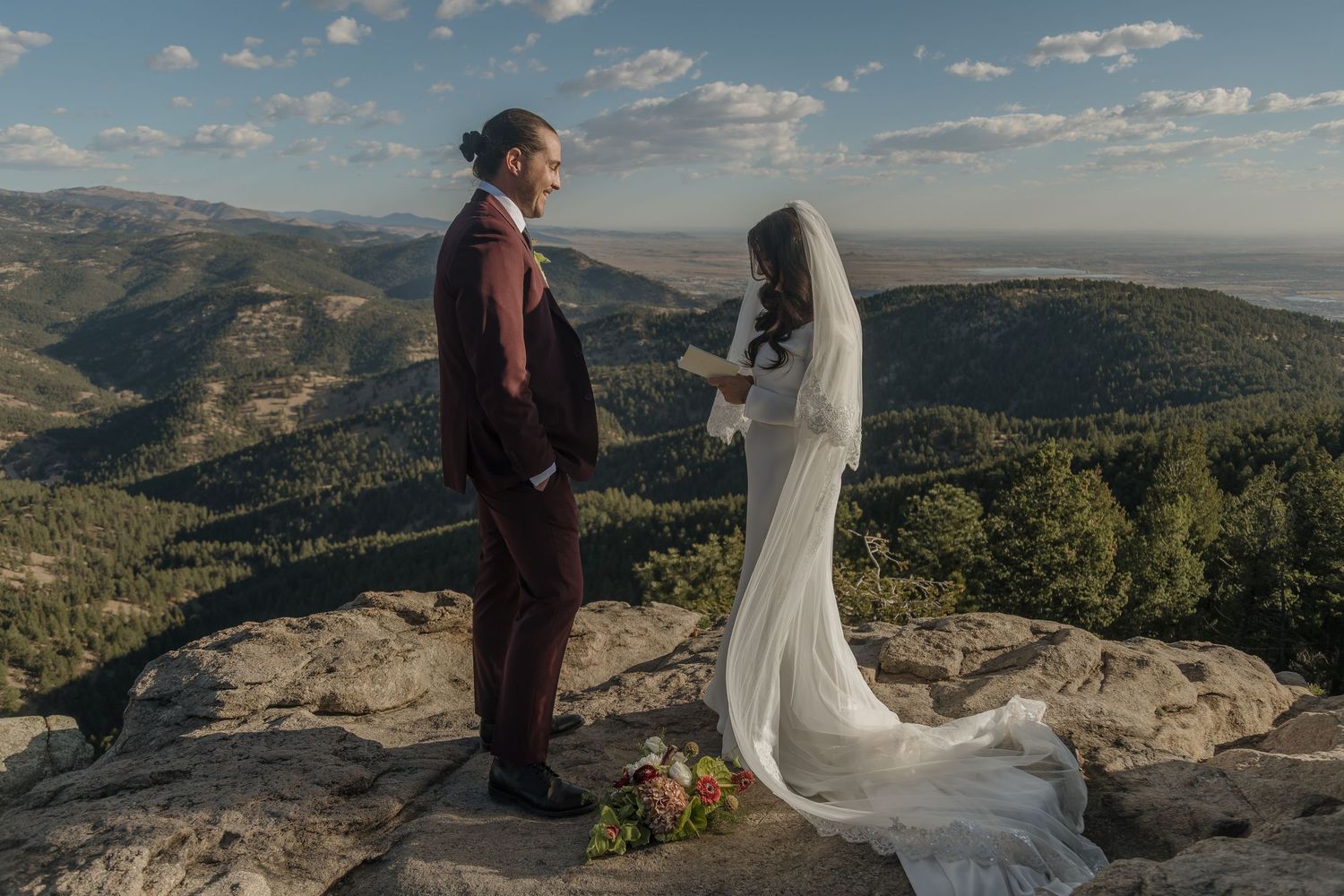 Bride in white gown and groom in burgundy suit exchanging vows on a dramatic mountain cliff.