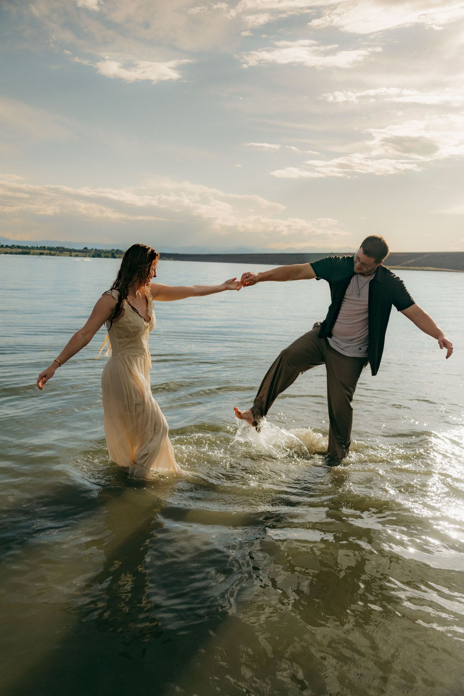 Couple laughing and splashing through shallow water at sunset during romantic beach engagement shoot.
