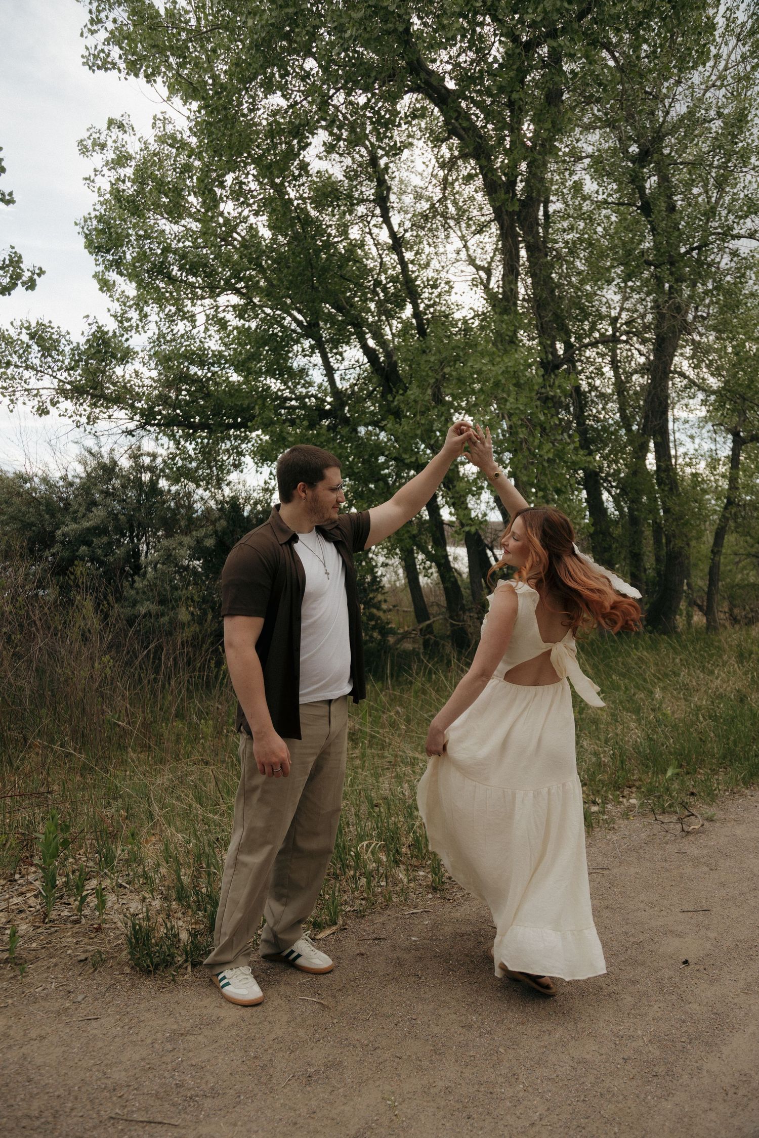 Man spinning woman in white dress on a dirt path surrounded by lush green trees outdoors.