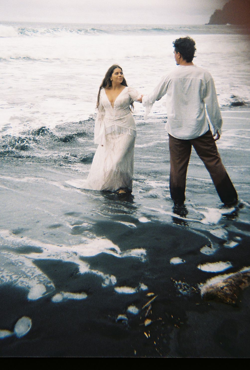 Couple holding hands in ocean waves, woman in white lace dress, man in light shirt, romantic beach photo shoot.