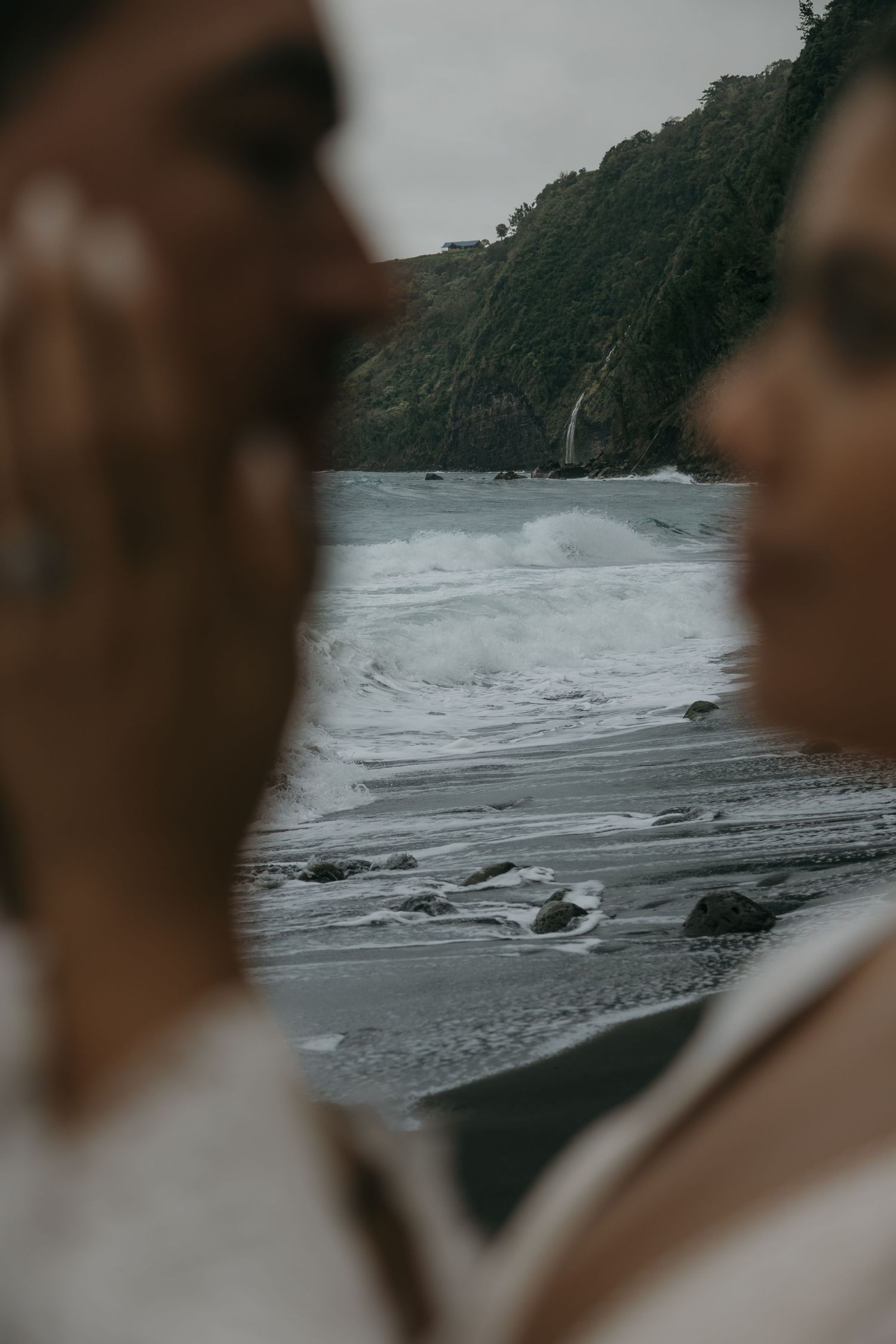 Artistic blurred shot of couple framing dramatic black sand beach with crashing waves and cliffs.