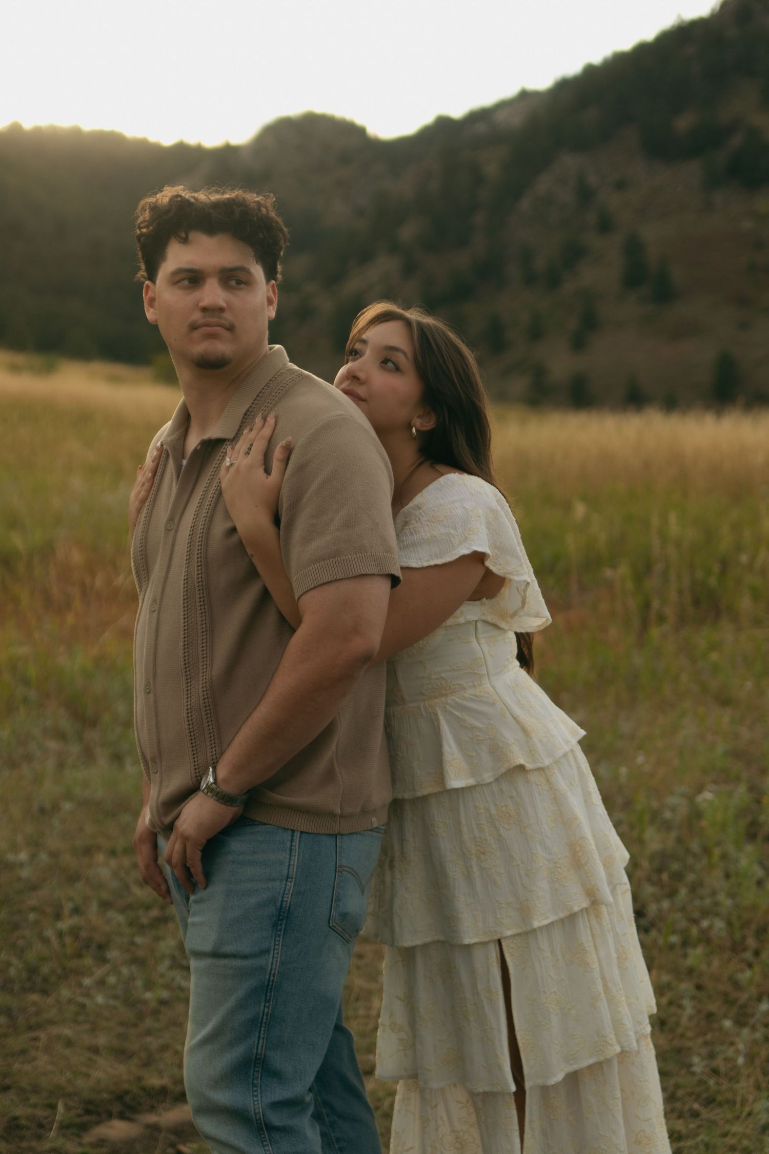 Couple poses at sunset in a mountain meadow, woman in white dress embraces man from behind.