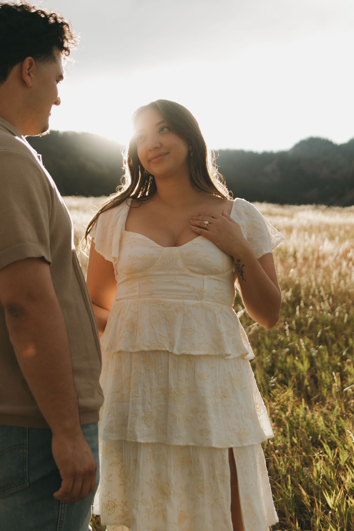 Woman in white tiered dress gazes at man in warm sunset light in open field.