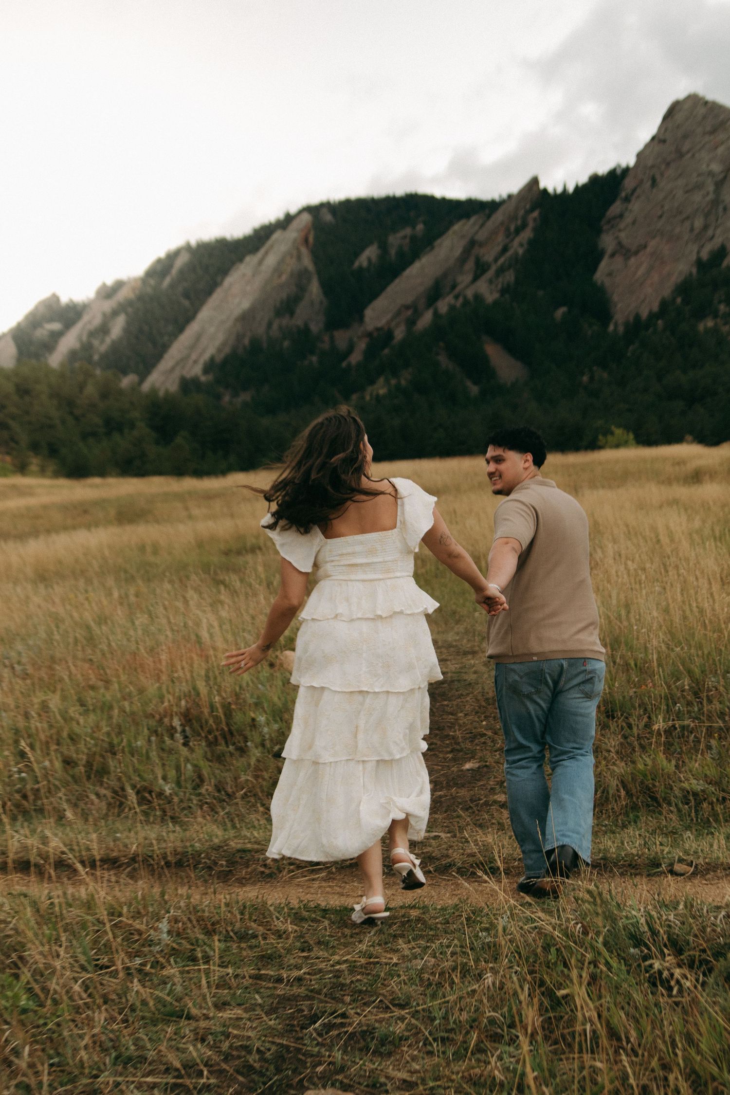 Couple holding hands walking through golden meadow with dramatic Flatirons rock formations in background.