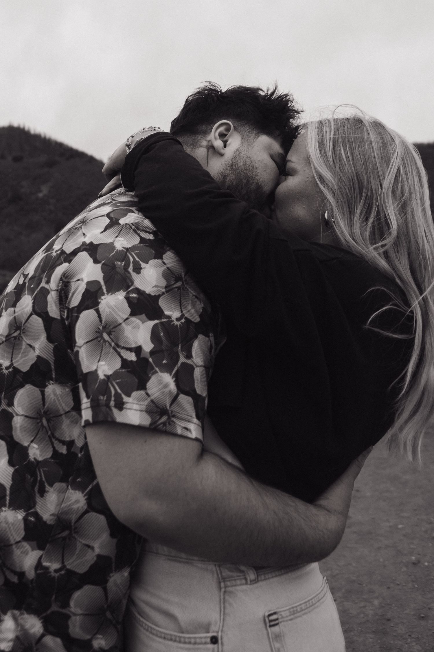 Black and white photo of a couple sharing a passionate kiss outdoors, woman with blonde hair embracing man in floral shirt.