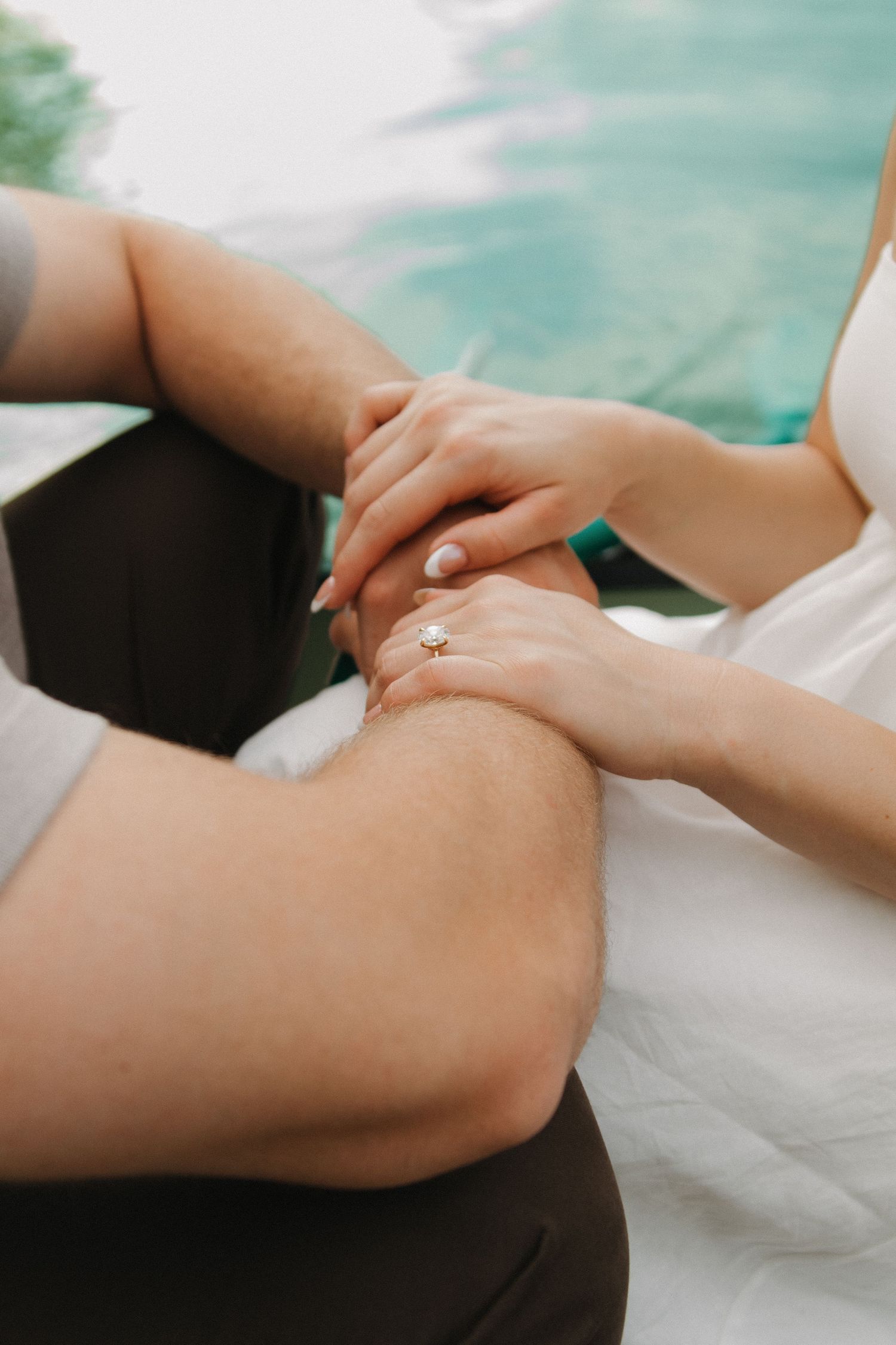 Close-up of a couple holding hands, woman wearing a diamond engagement ring, sitting near a turquoise pool.