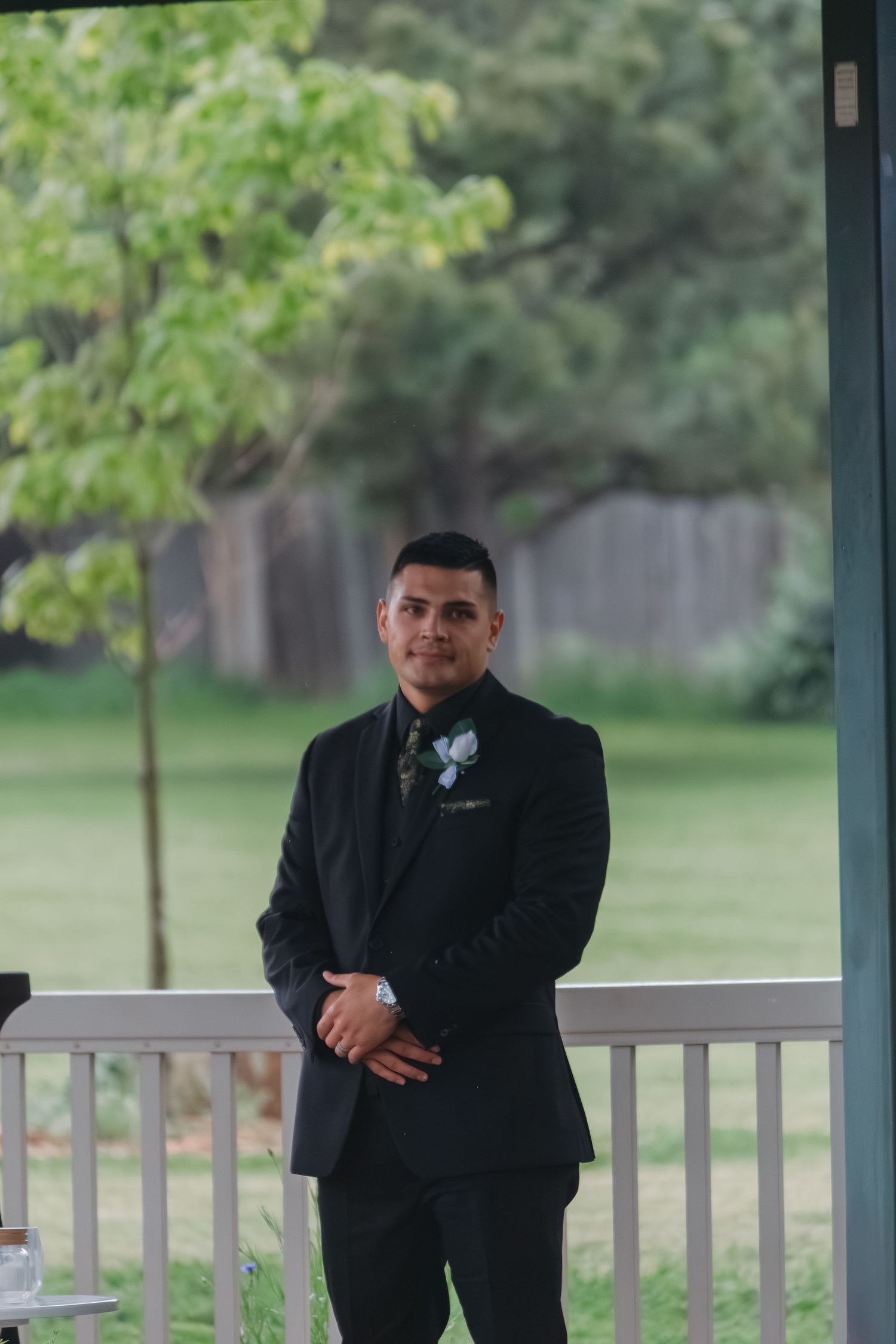 A person in a black suit stands on a covered porch with trees in the background.