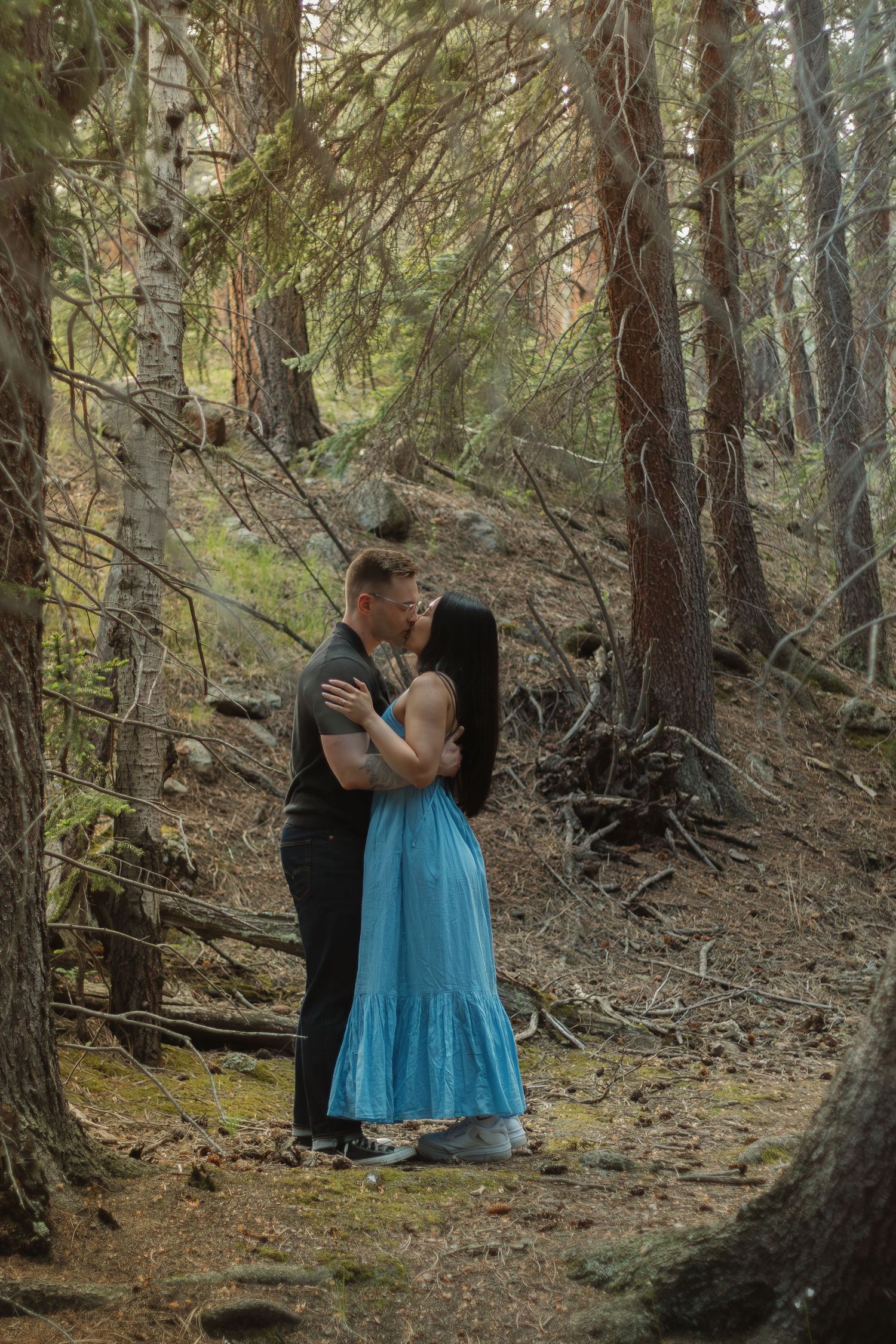A couple shares an intimate moment on a wooded hillside wearing formal attire.