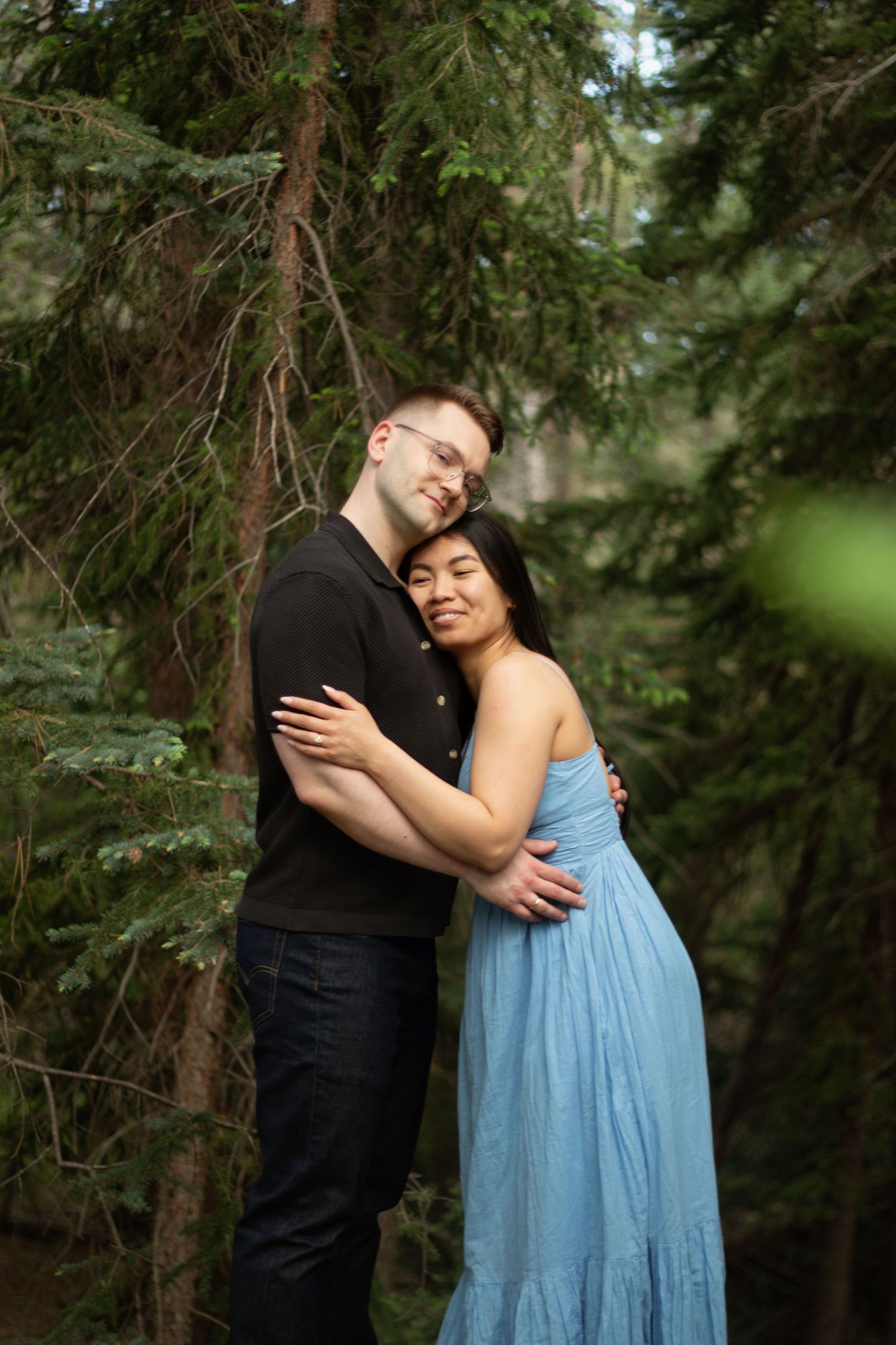 Two people embrace lovingly among evergreen trees in a romantic forest photoshoot.