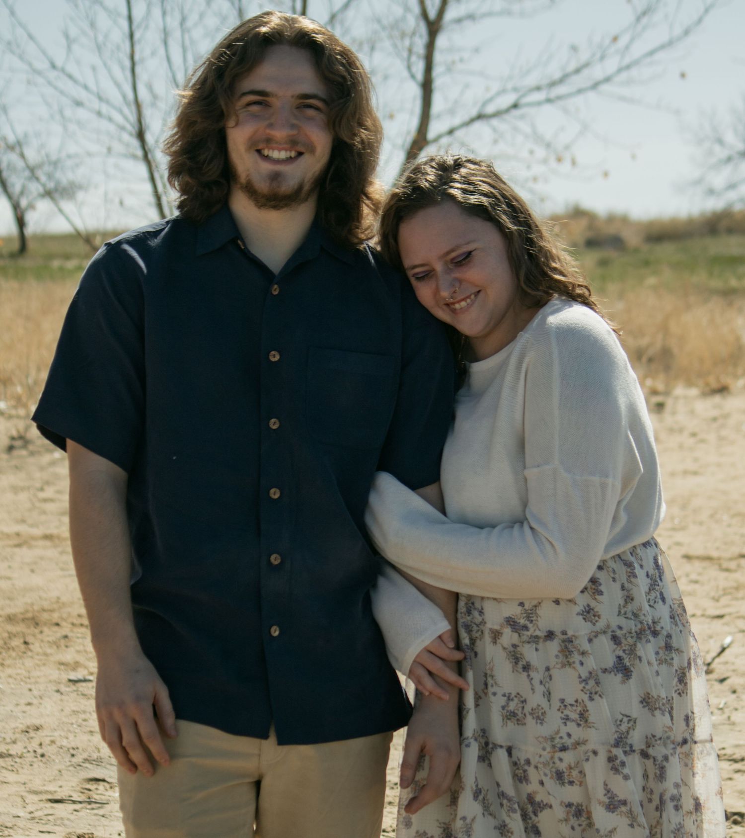 A happy couple standing together in casual attire against a rural backdrop.