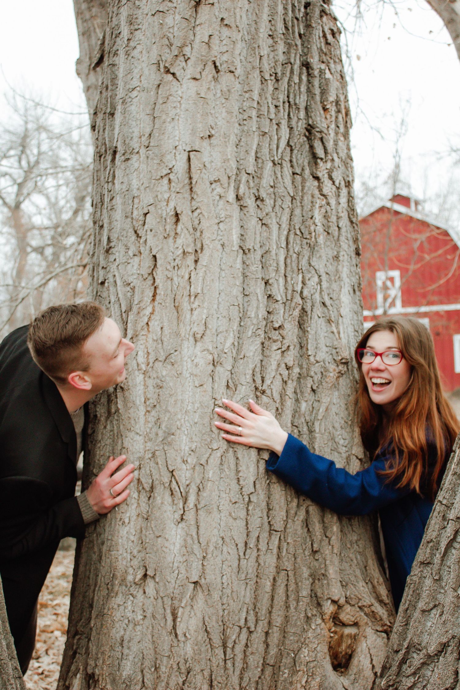 A playful moment captured as a couple peeks around opposite sides of a large tree trunk.