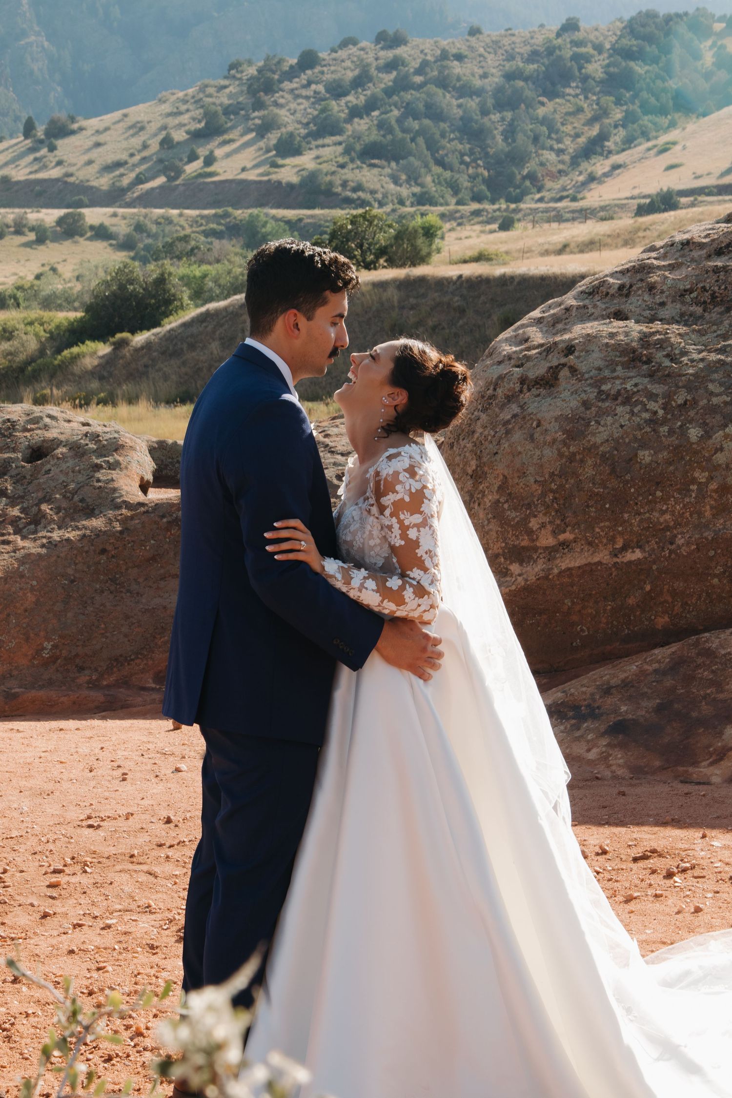 A couple in formal wedding attire embraces on red rocky terrain with mountains in the distance.