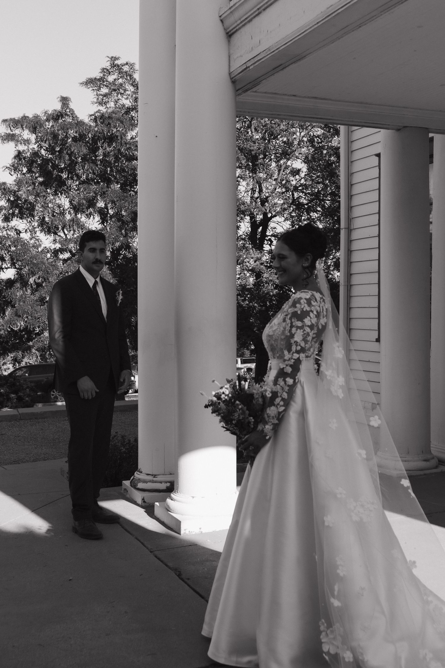 Elegant wedding couple poses between white columns in black and white photography.