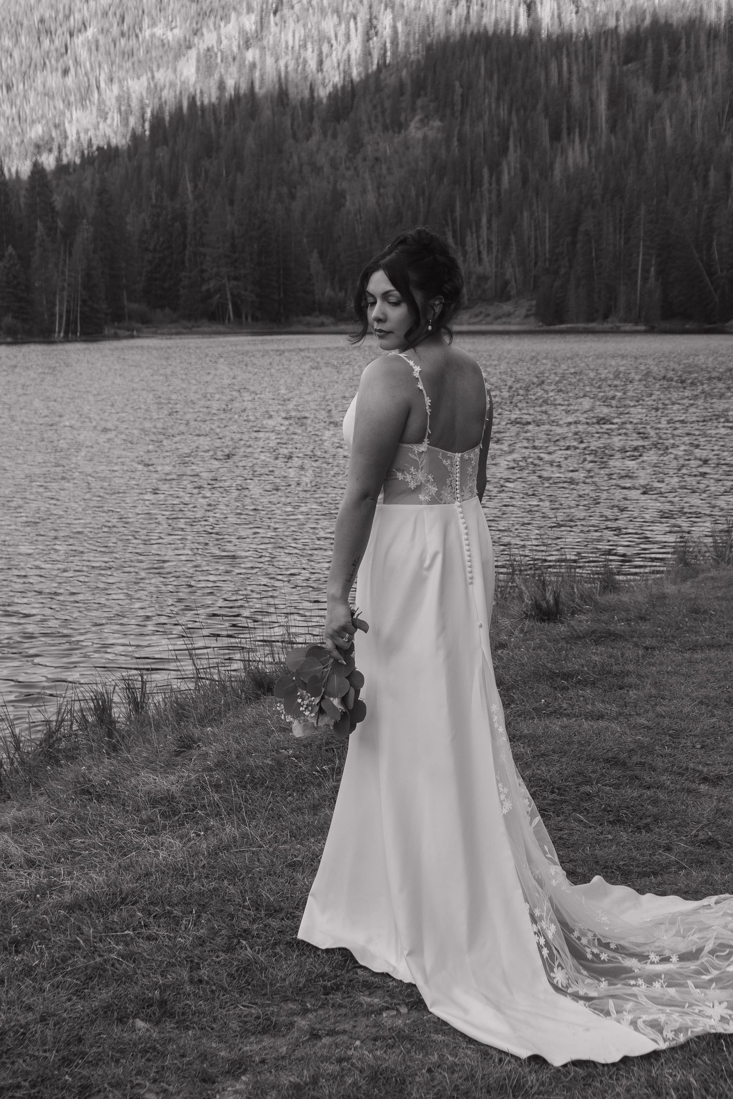 Flowing white wedding dress cascades across green grass near a serene mountain lake.