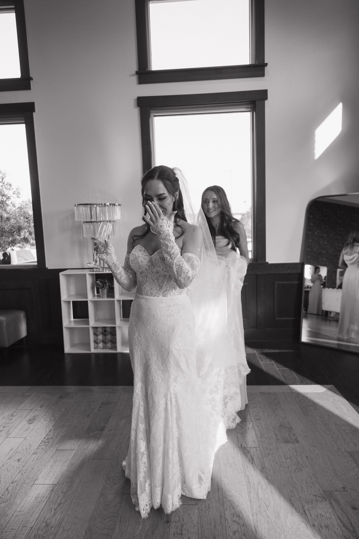Emotional bride in lace wedding gown wipes tears while bridesmaid holds her veil in black and white photo.