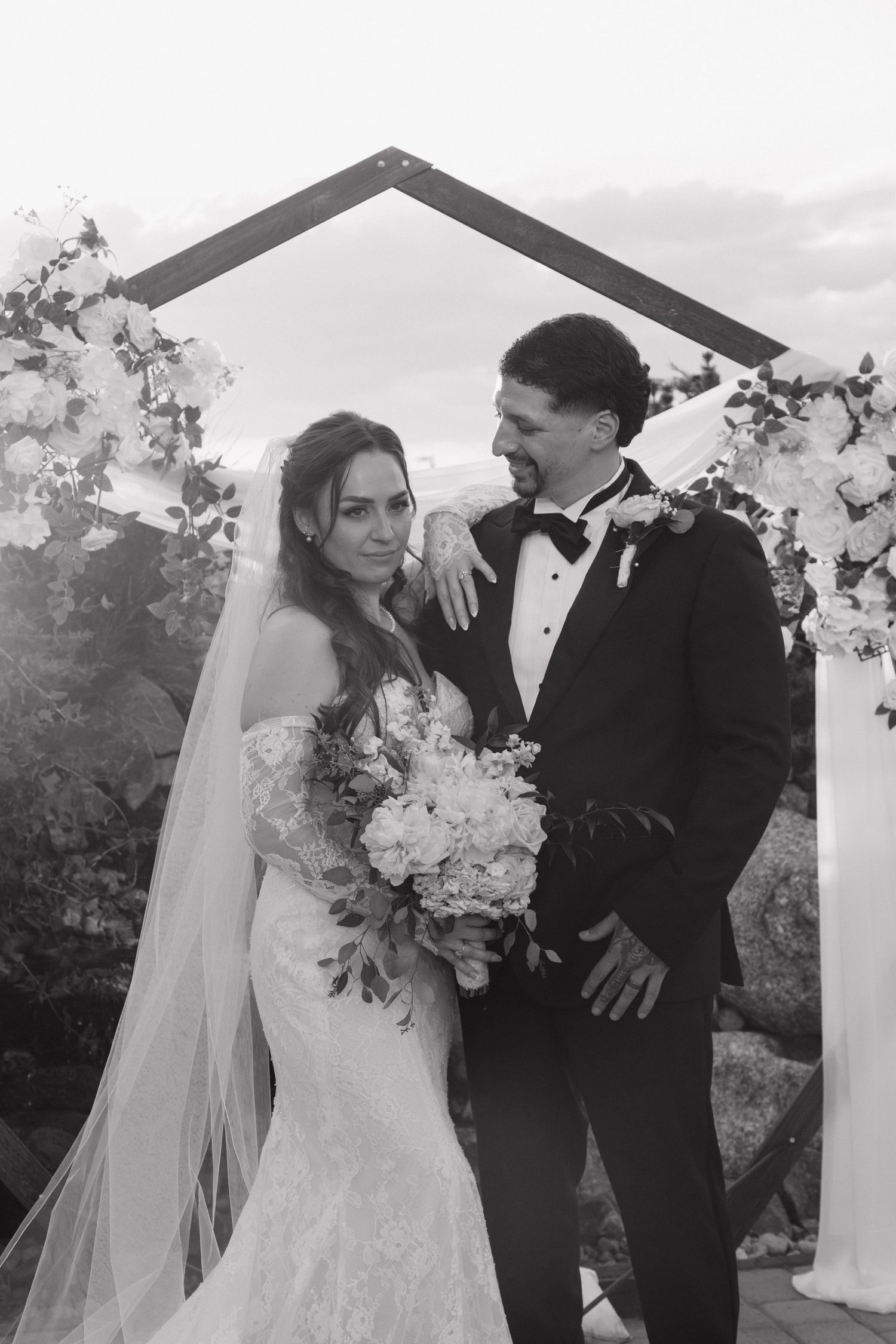 Black and white photo of a bride and groom posing under a floral arch, bride holding a bouquet in a wedding gown.