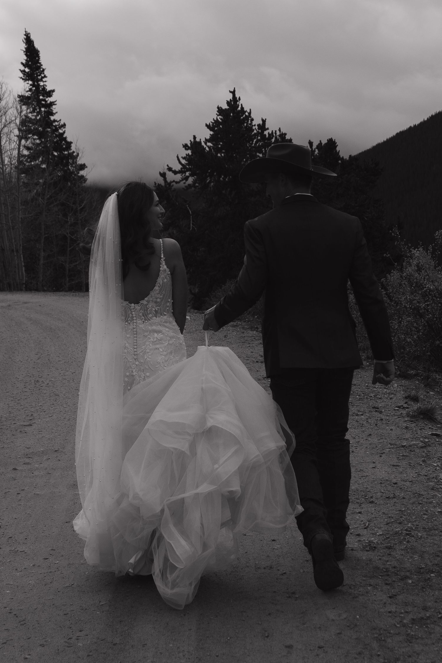 A couple walks through a snowy mountain landscape with evergreen trees in the background.