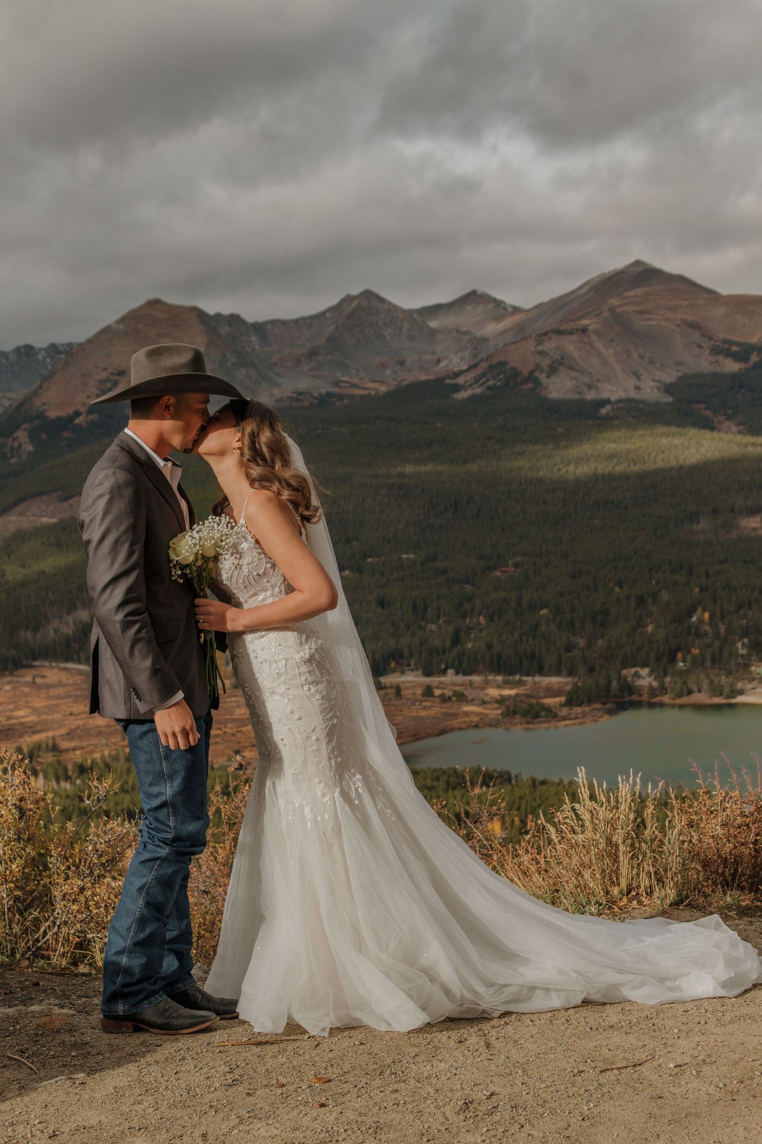 Newlyweds share romantic moments on a mountain overlook with a scenic lake and peaks in the background.