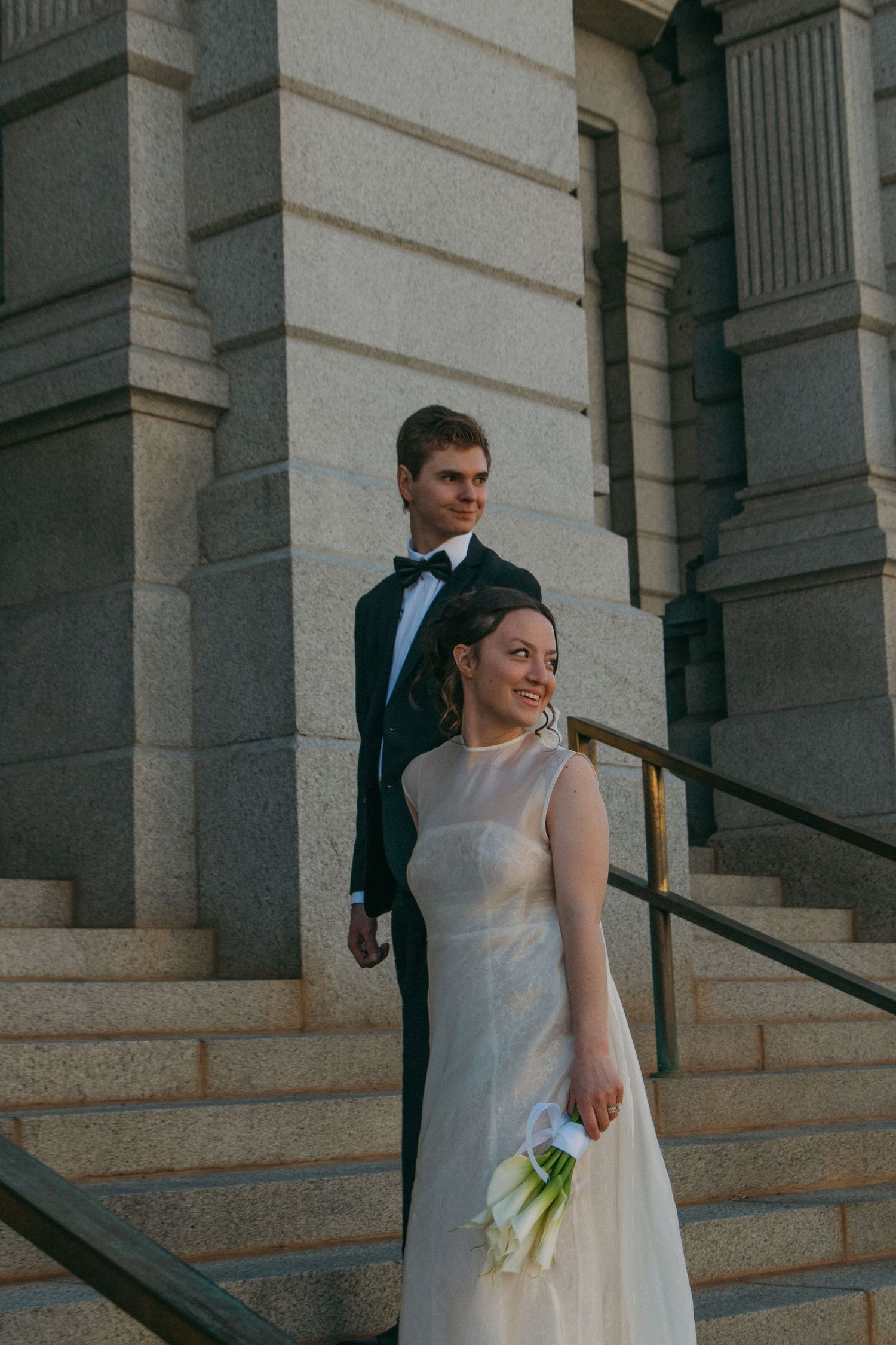 A wedding couple descends grand stone steps of a historic building with classical columns.