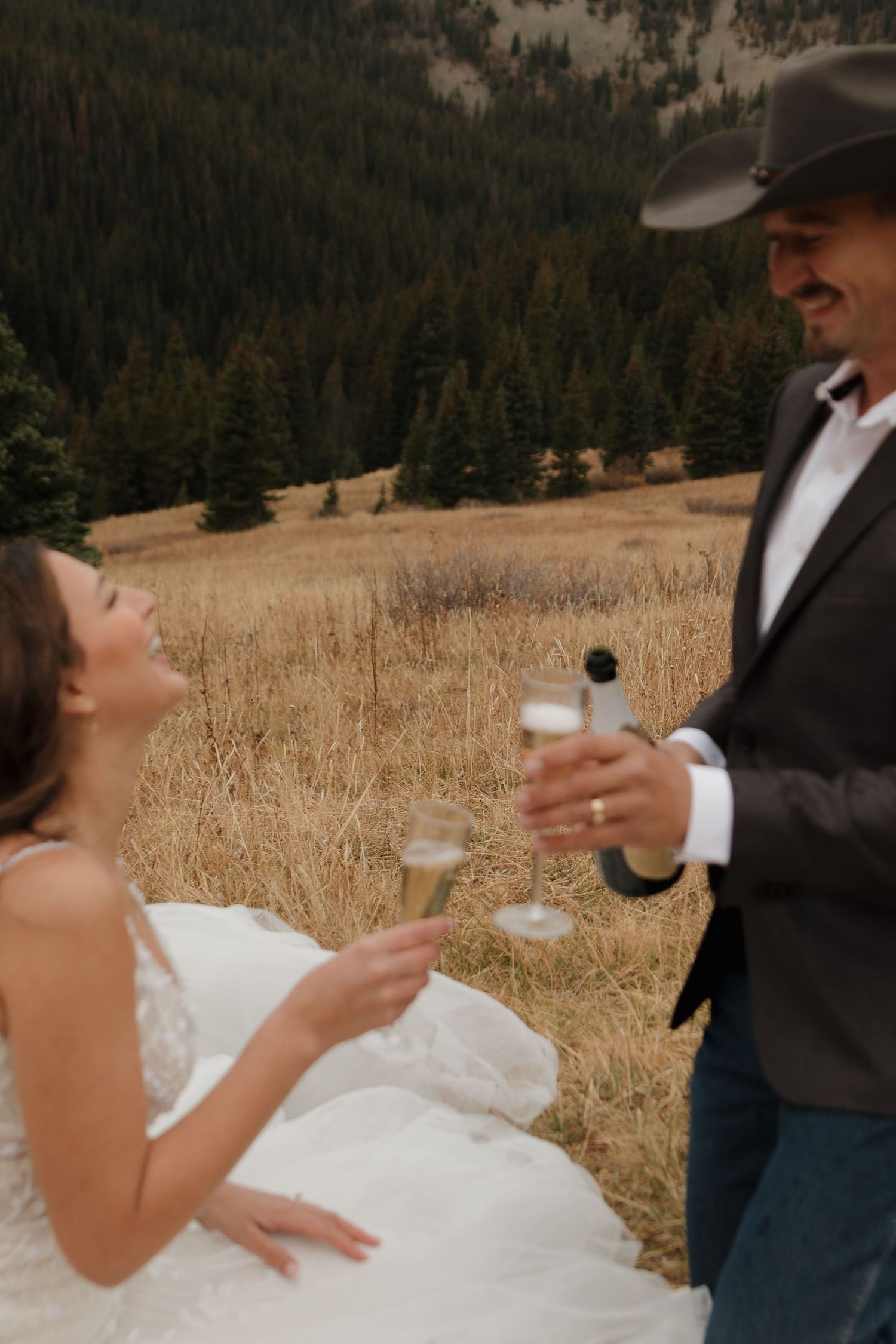 Bride and groom toast with champagne glasses on a scenic mountain hillside.