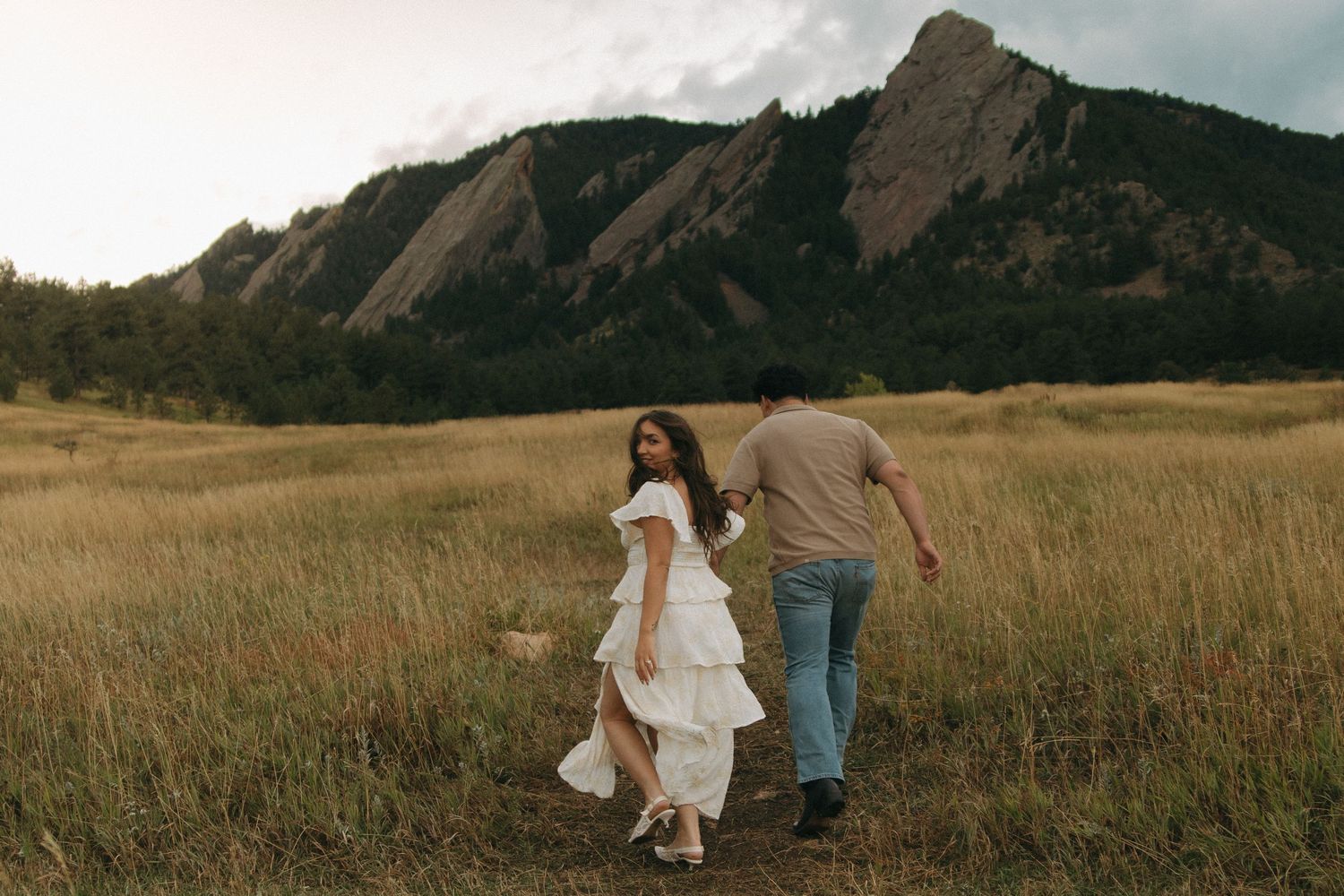 A couple walks through a golden meadow with dramatic mountain peaks in the background at sunset.