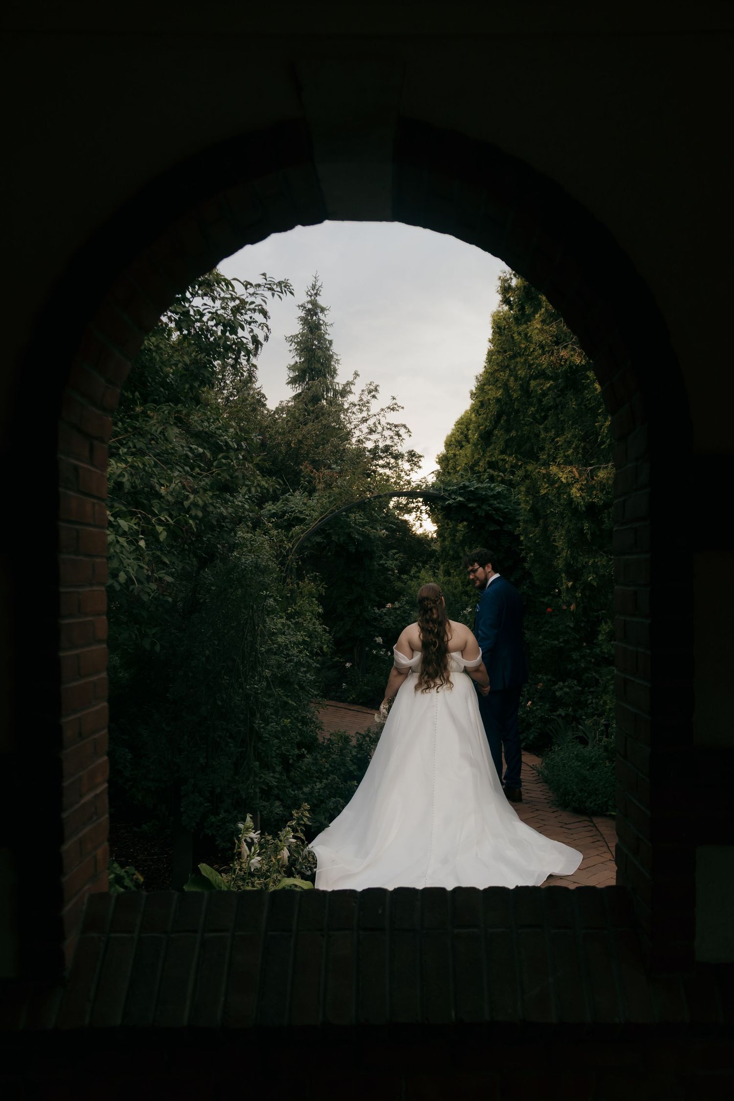 Bride and groom framed by arched brick doorway in lush garden setting on their wedding day.