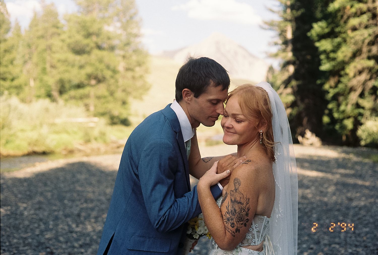 Groom tenderly kisses bride's forehead as she smiles, showcasing her floral arm tattoo outdoors.