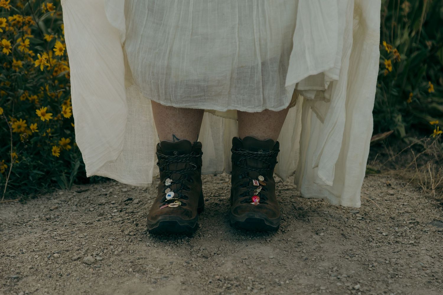 Bride wearing hiking boots with floral pins under a flowing white dress on a dirt trail.