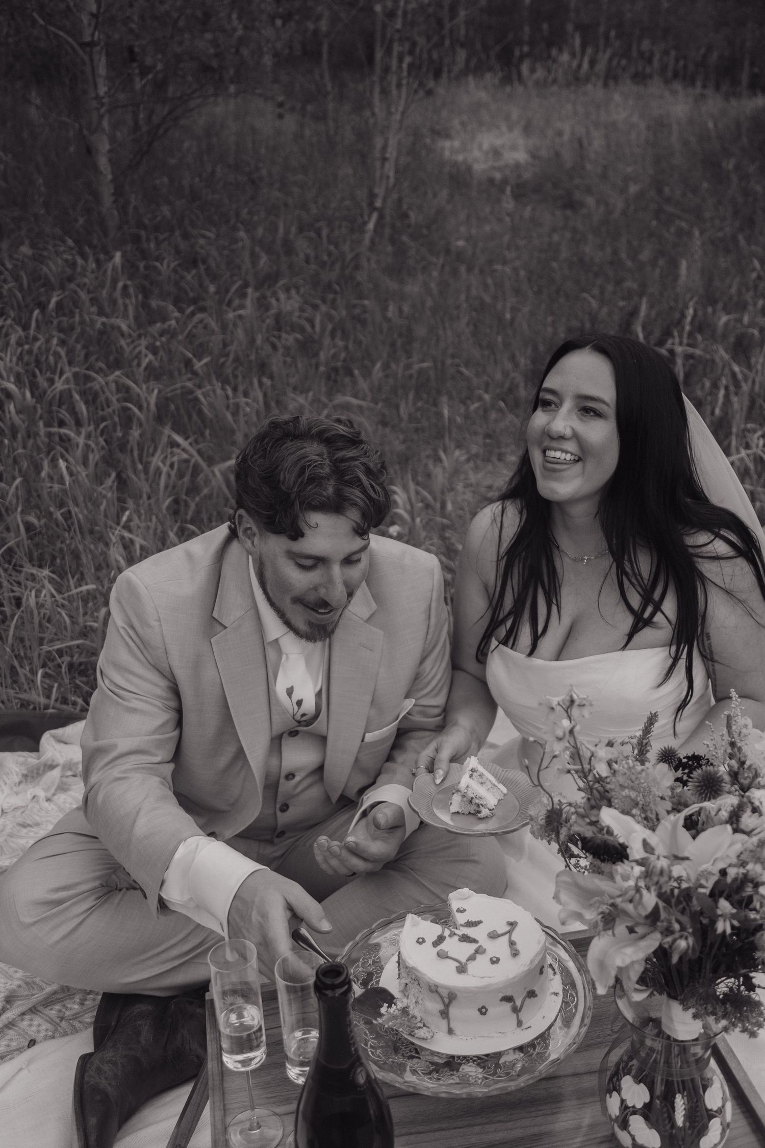 Black and white photo of laughing bride and groom cutting their wedding cake during forest picnic.