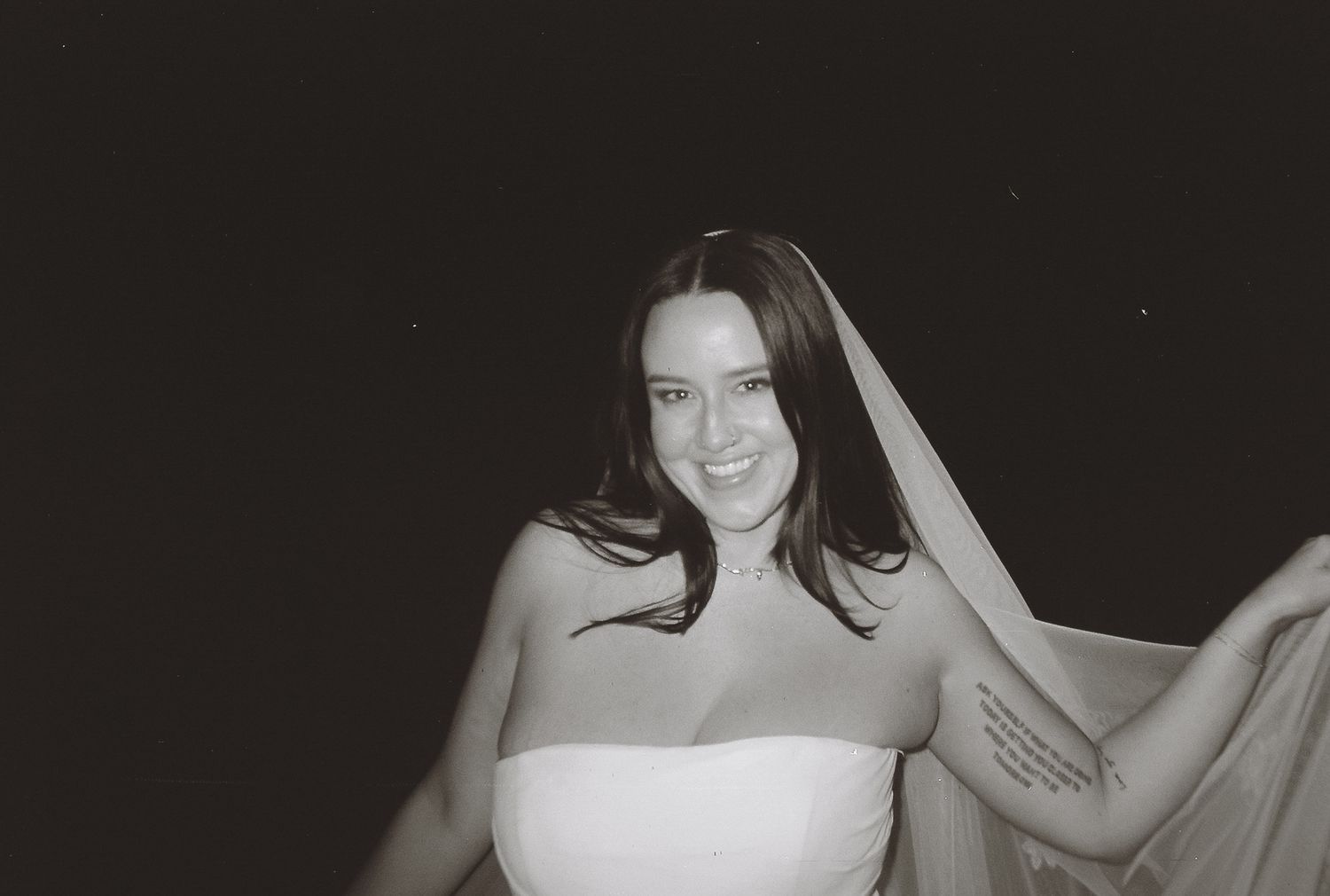 Black and white film photo of a smiling bride in a strapless gown holding her veil against a dark background.