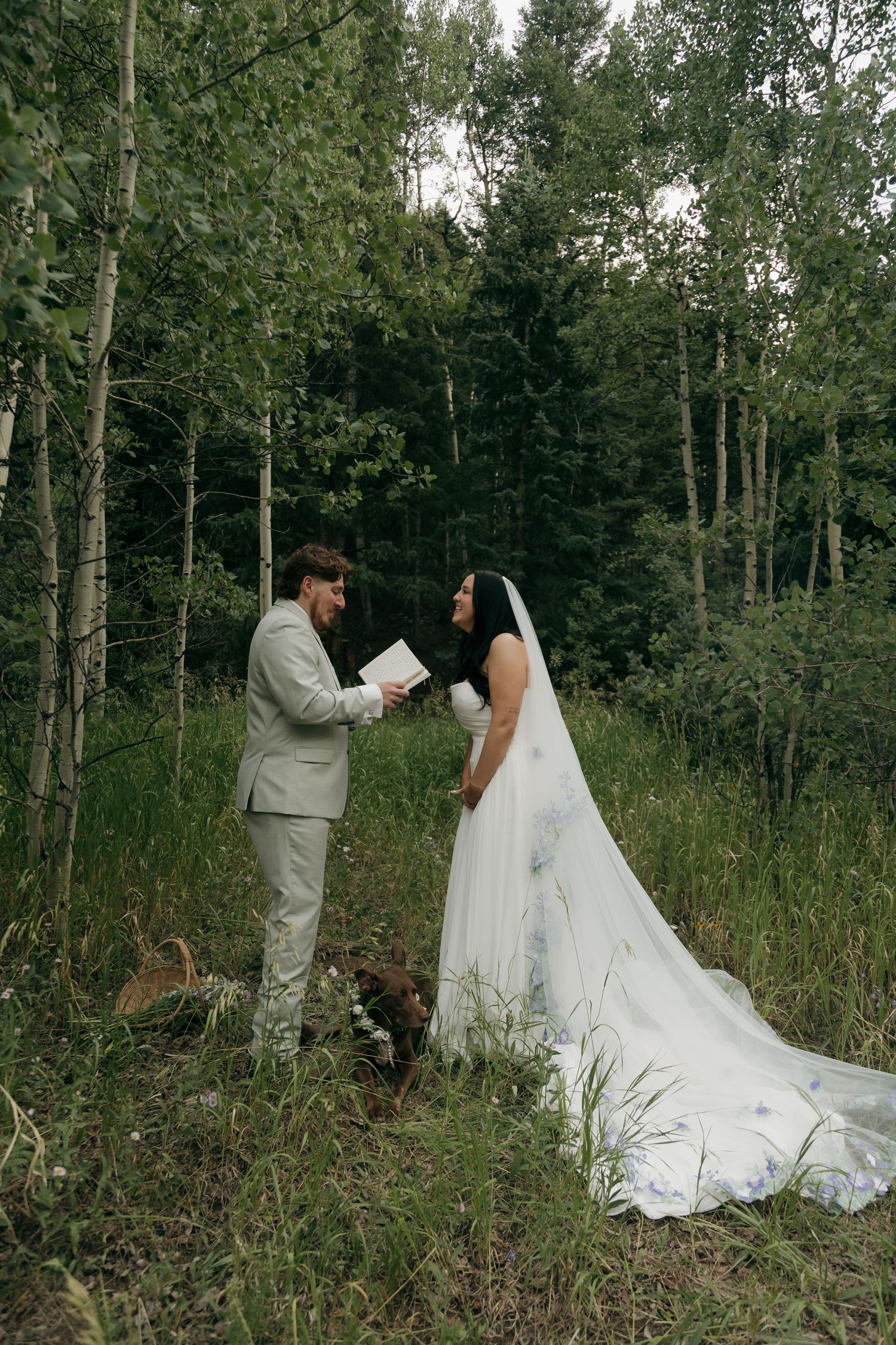 Bride and groom exchange vows in a lush forest clearing, she in a floral-embroidered gown with long veil.