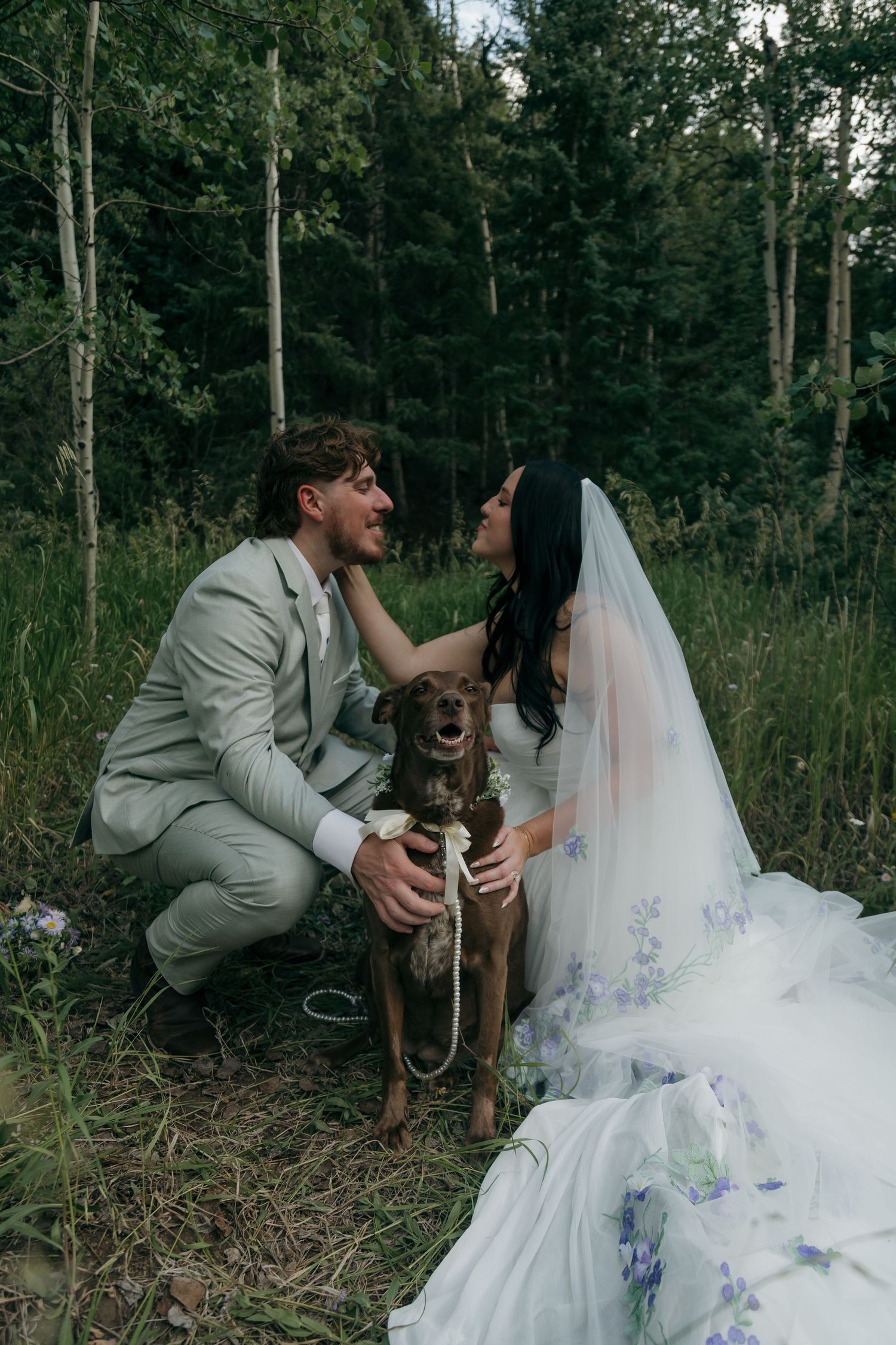 Bride and groom crouch together smiling at their brown dog during an outdoor forest elopement.